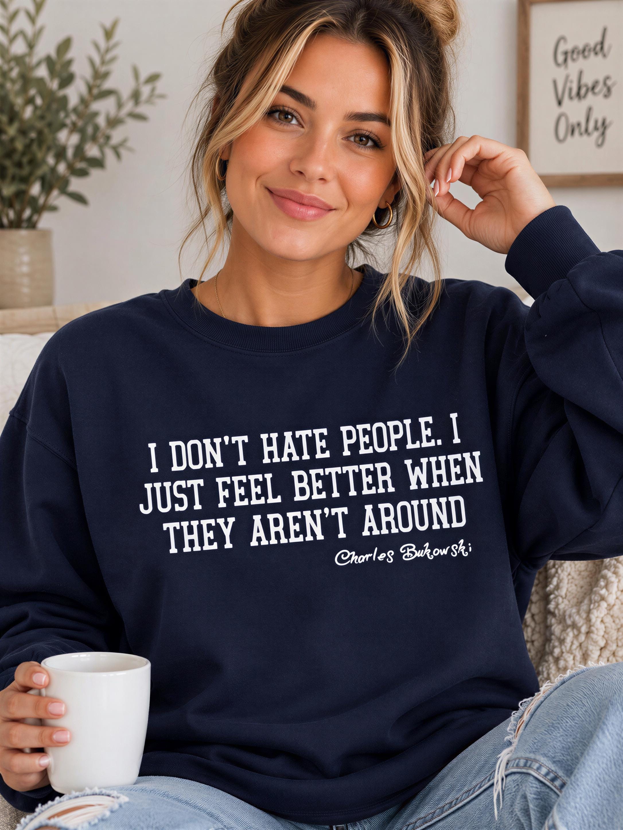 A woman wearing a navy blue sweatshirt with a humorous text printed on it, sitting on a couch and holding a white mug.