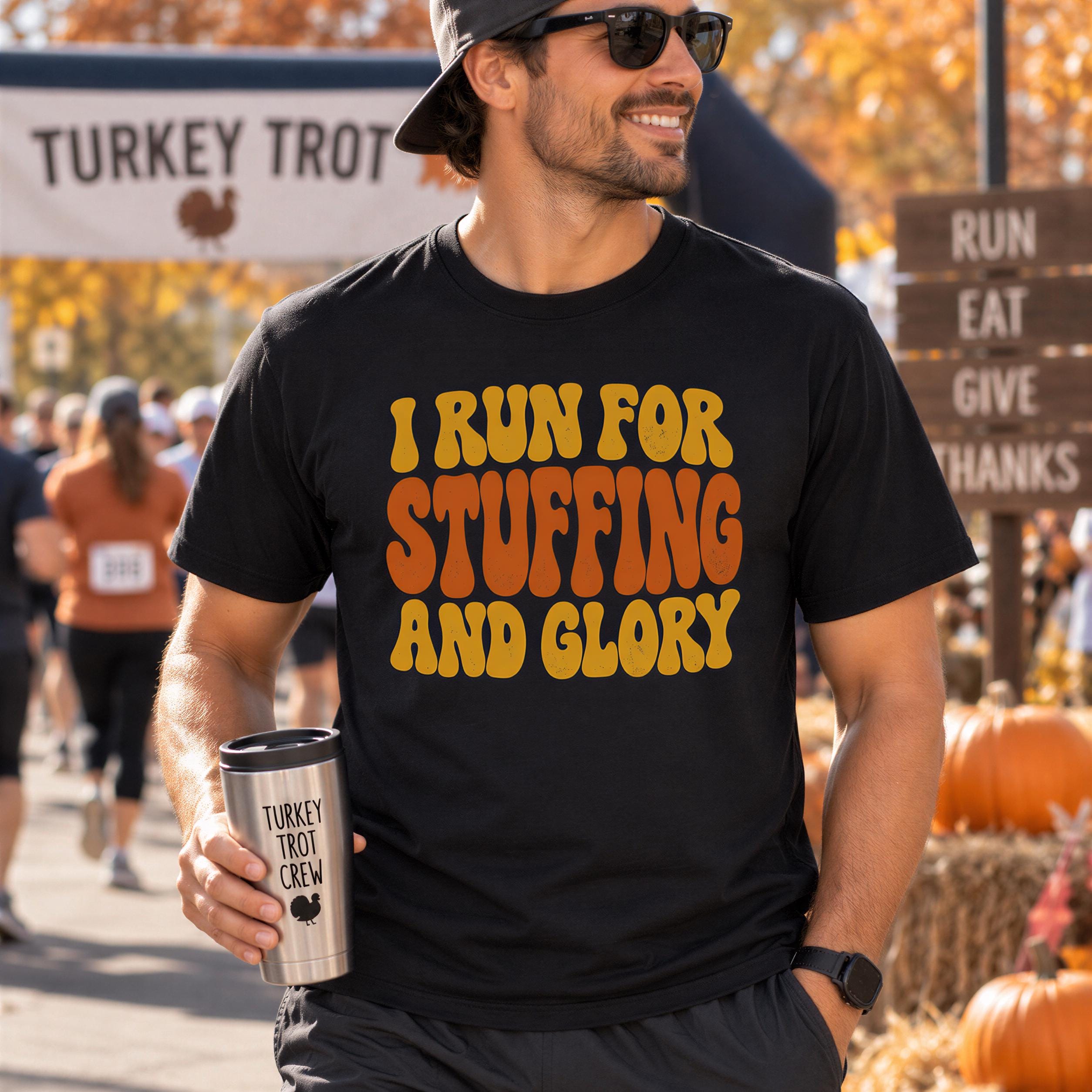 A man wearing a black t-shirt with a humorous text printed on it, smiling and holding a turkey trot crew mug, with a sign for a turkey trot event in the background.
