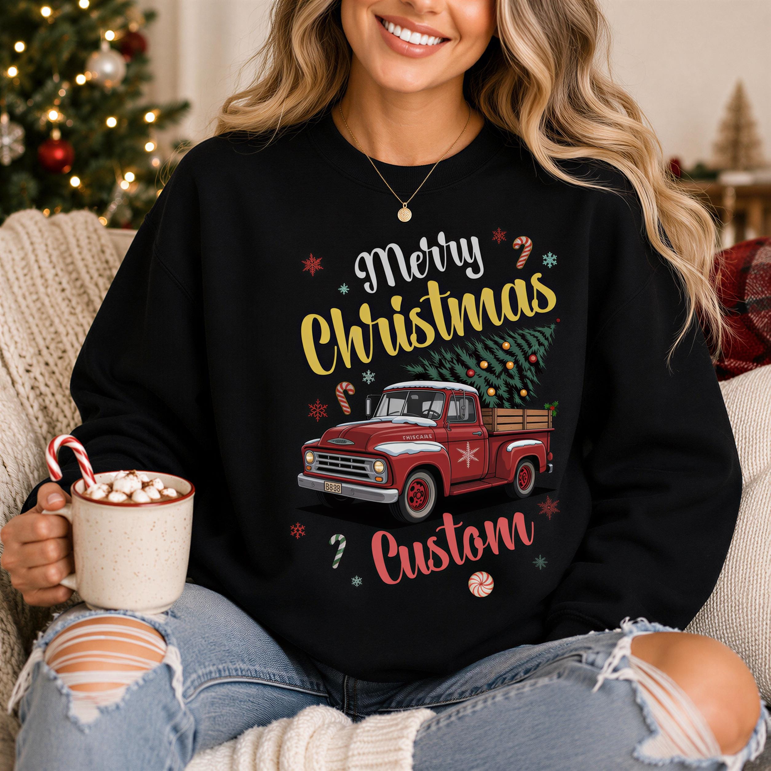 A woman wearing a black sweatshirt with a Christmas-themed design, including a red truck and Christmas decorations, is sitting on a couch and holding a mug of hot chocolate.