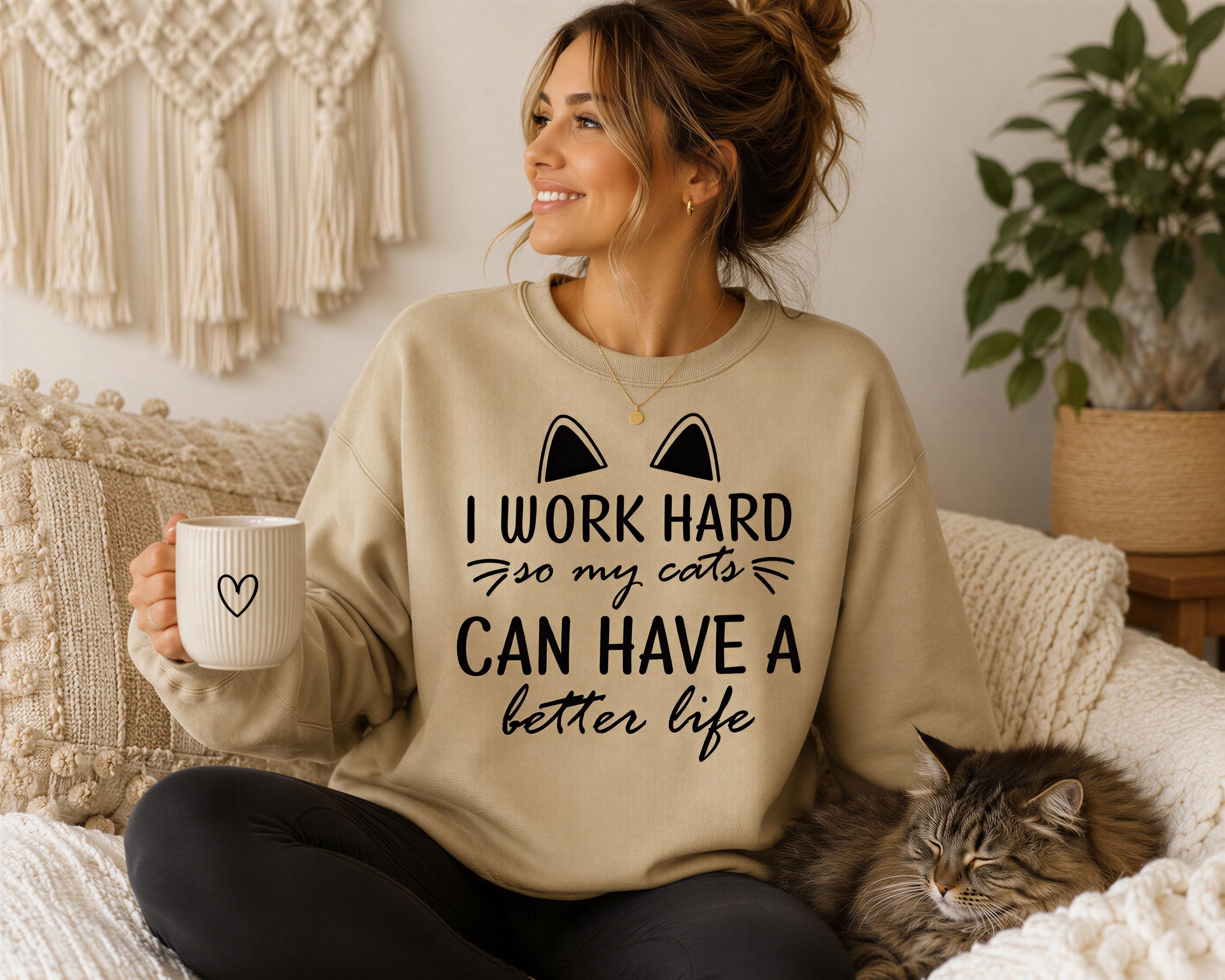 A woman sitting on a couch, wearing a sweatshirt with the text "I WORK HARD so my cats can have a better life" and holding a coffee mug. A cat is sleeping next to her.