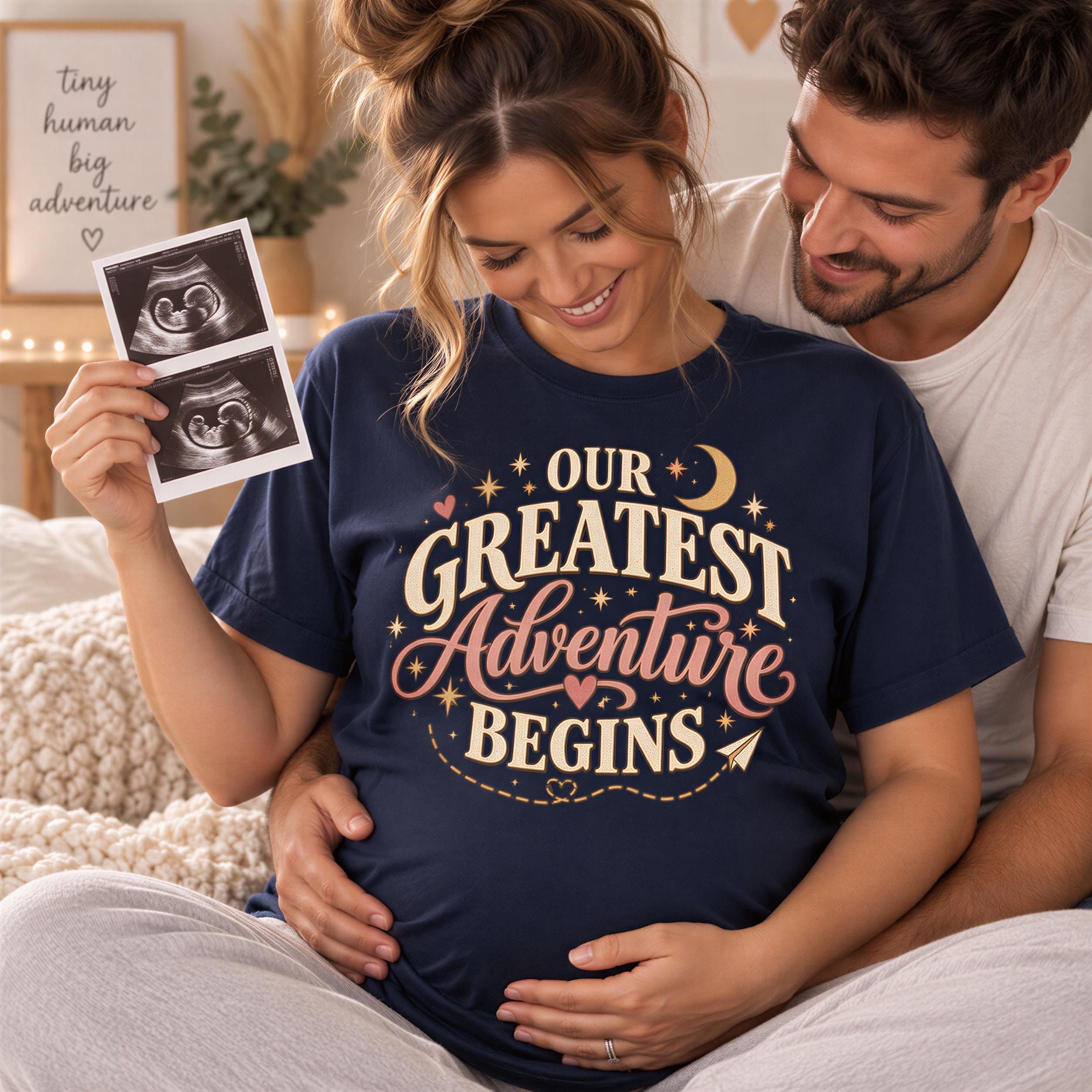 A pregnant woman wearing a blue t-shirt with the text "Our Greatest Adventure Begins" is shown holding an ultrasound image, with a smiling man standing next to her.