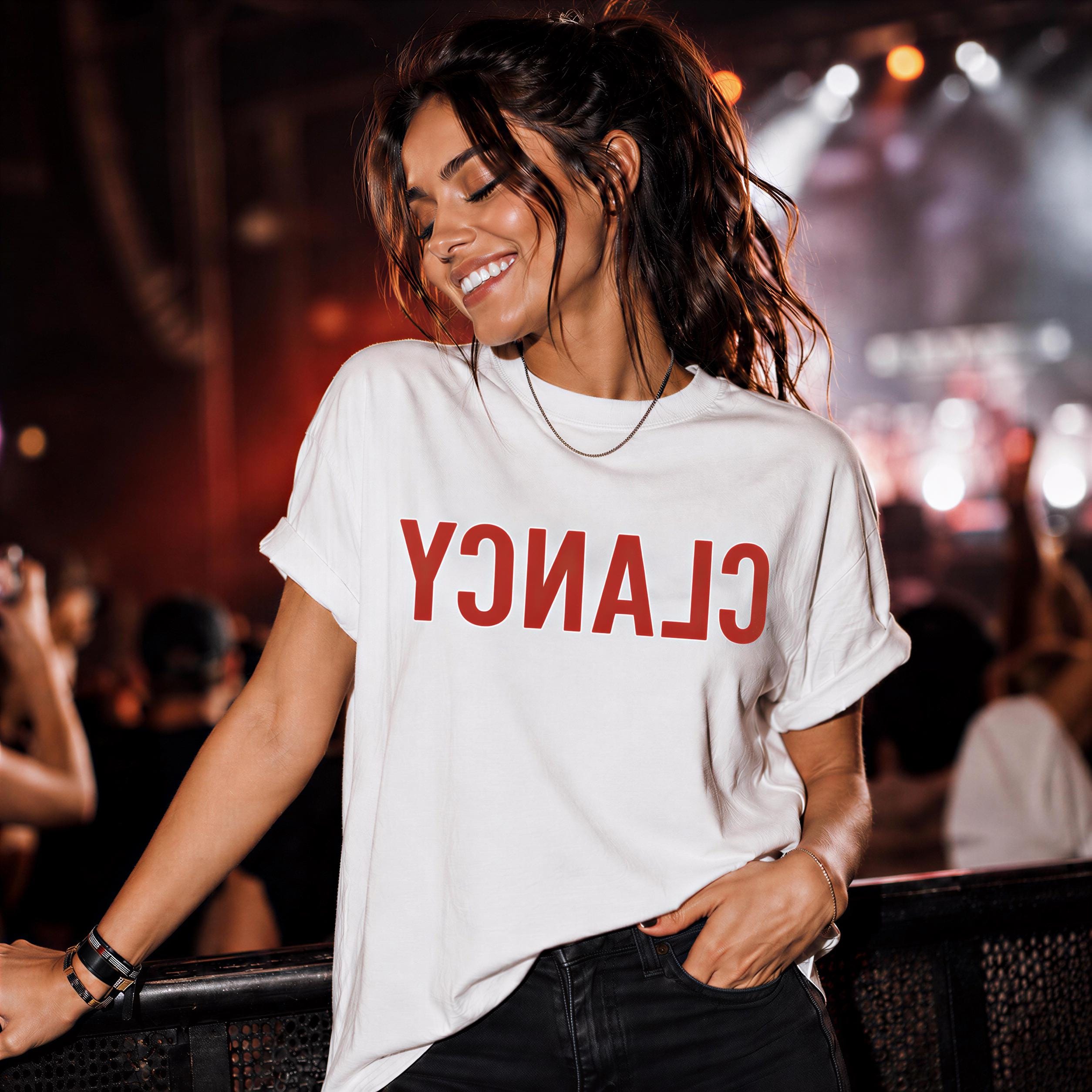 A woman wearing a white t-shirt with the word "CLAN" on it is smiling and leaning against a barrier at a concert.