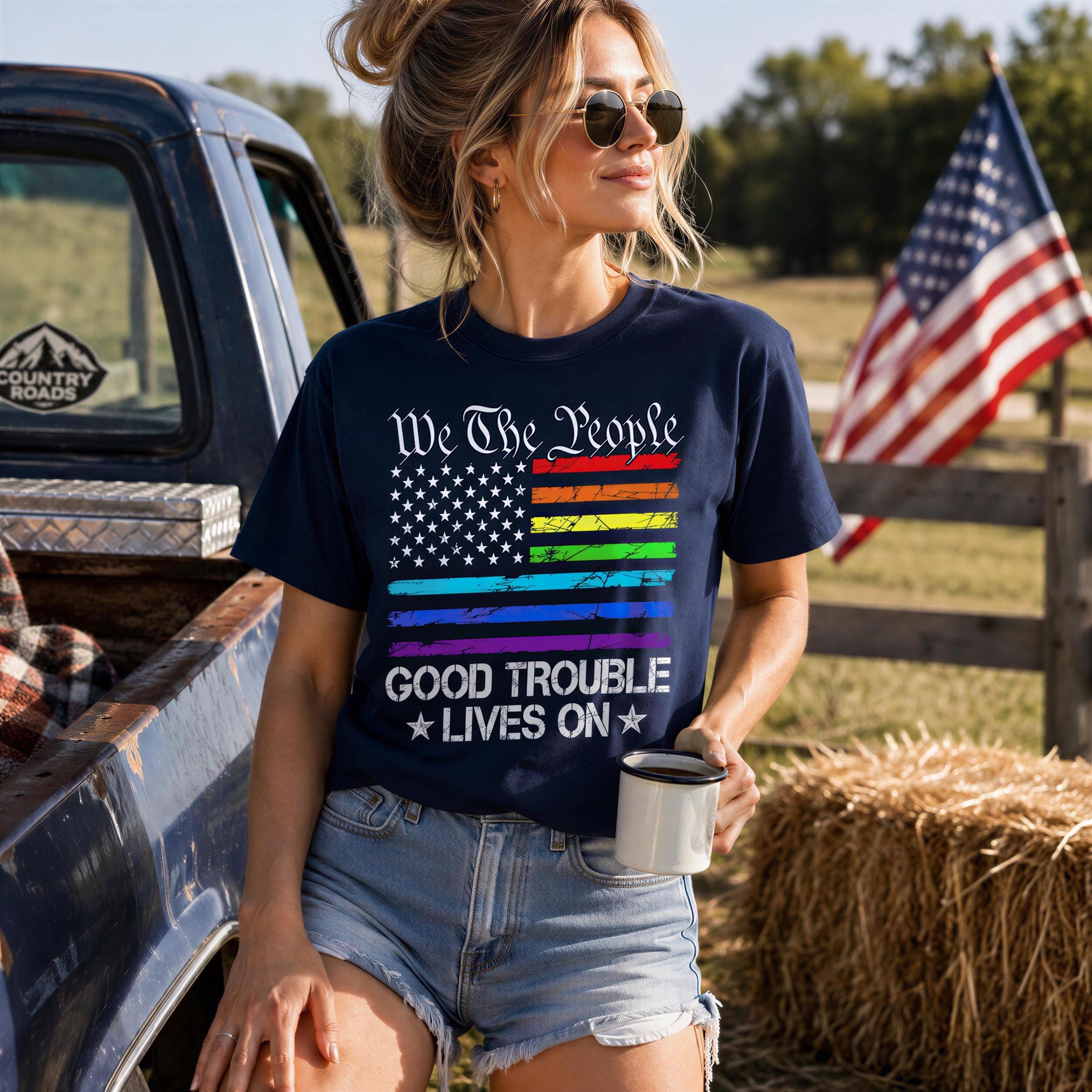 A woman wearing a blue t-shirt with a rainbow flag design and the text "We the People" stands next to a blue pickup truck in a rural setting.