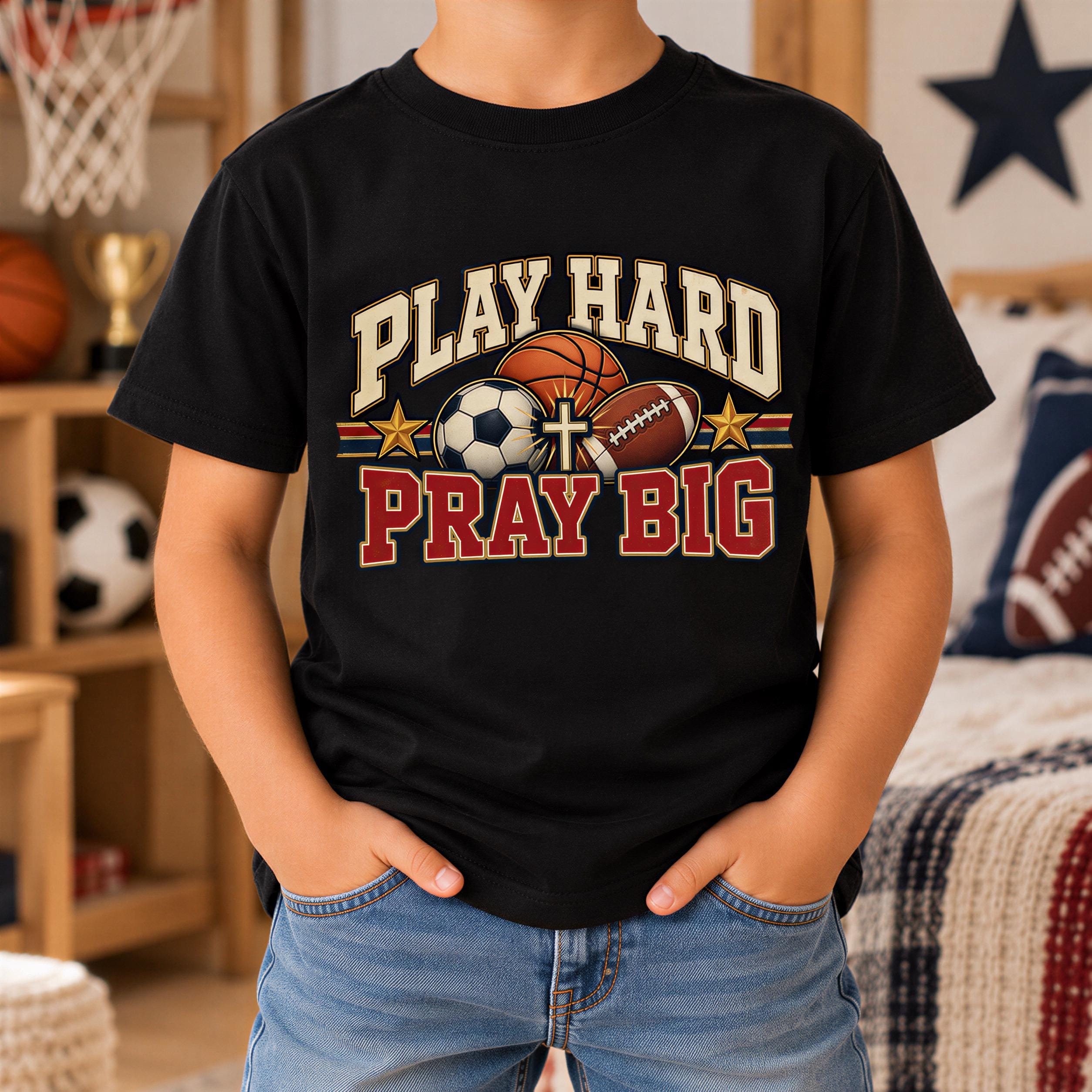 A person wearing a black t-shirt with a graphic design featuring sports balls and a cross, along with the text "PLAY HARD PRAY BIG".