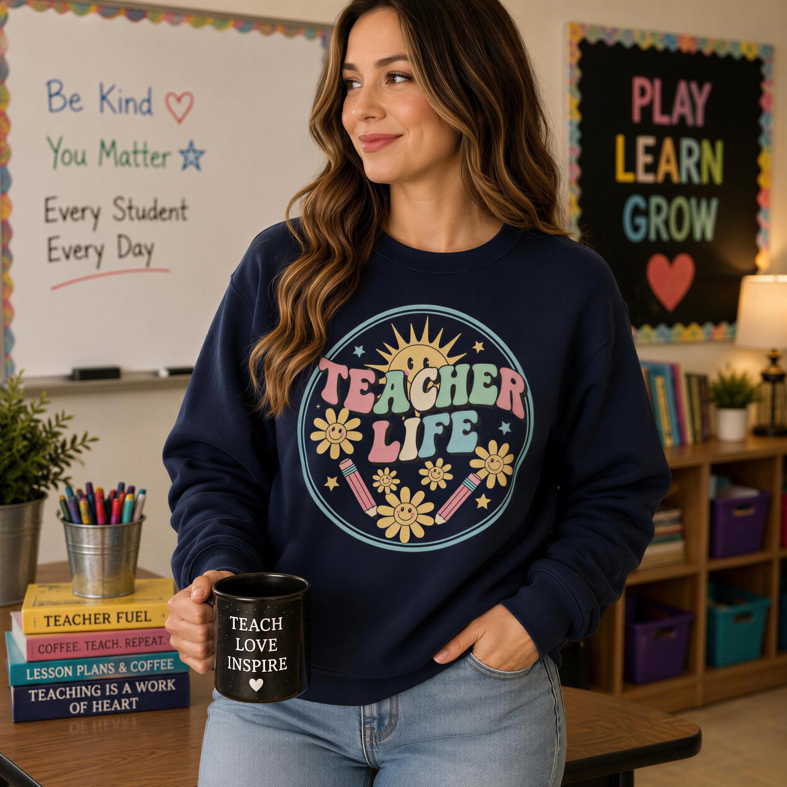 A woman wearing a blue sweatshirt with a colorful design stands in a classroom, holding a black mug with the text "TEACH LOVE INSPIRE" printed on it.