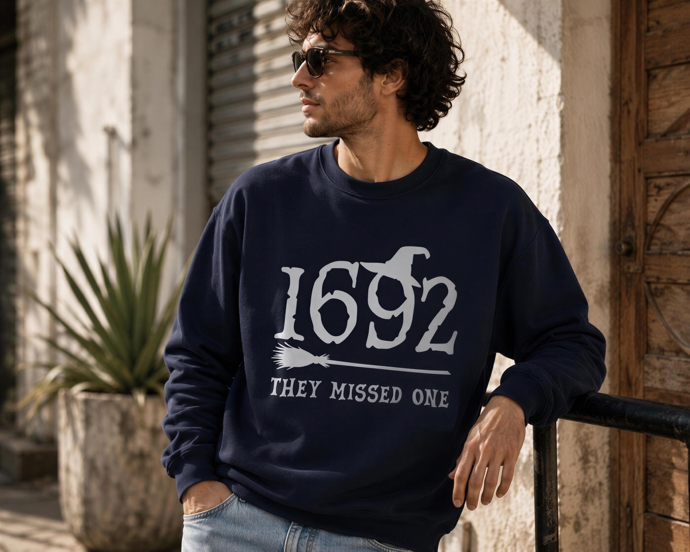 A man wearing a navy blue sweatshirt with the text "1692 THEY MISSED ONE" stands in front of a building.