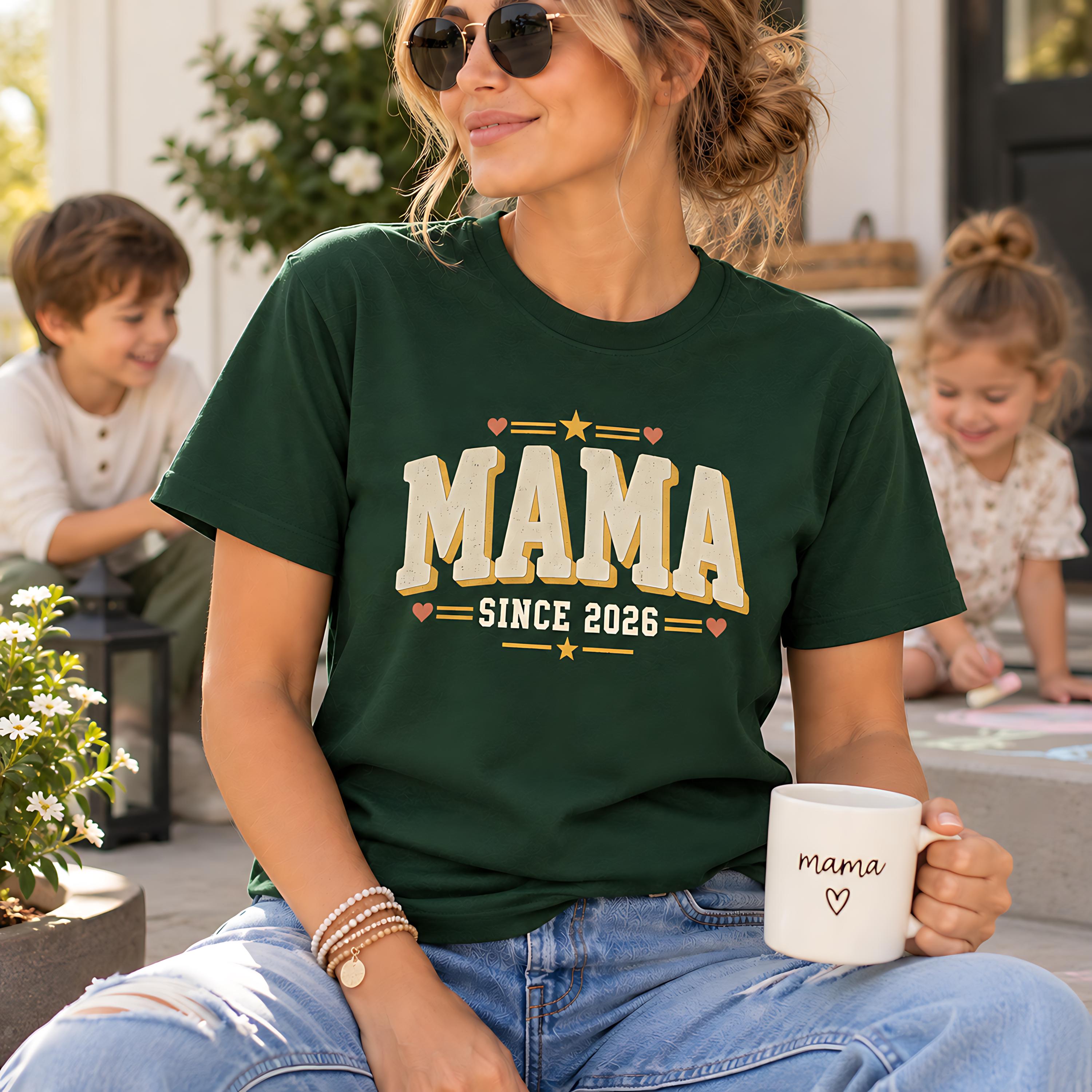 A woman wearing a green t-shirt with the text "MAMA SINCE 2026" is sitting on a porch, holding a white mug with the word "mama" printed on it. In the background, there are two children, one of whom is drawing on the steps.