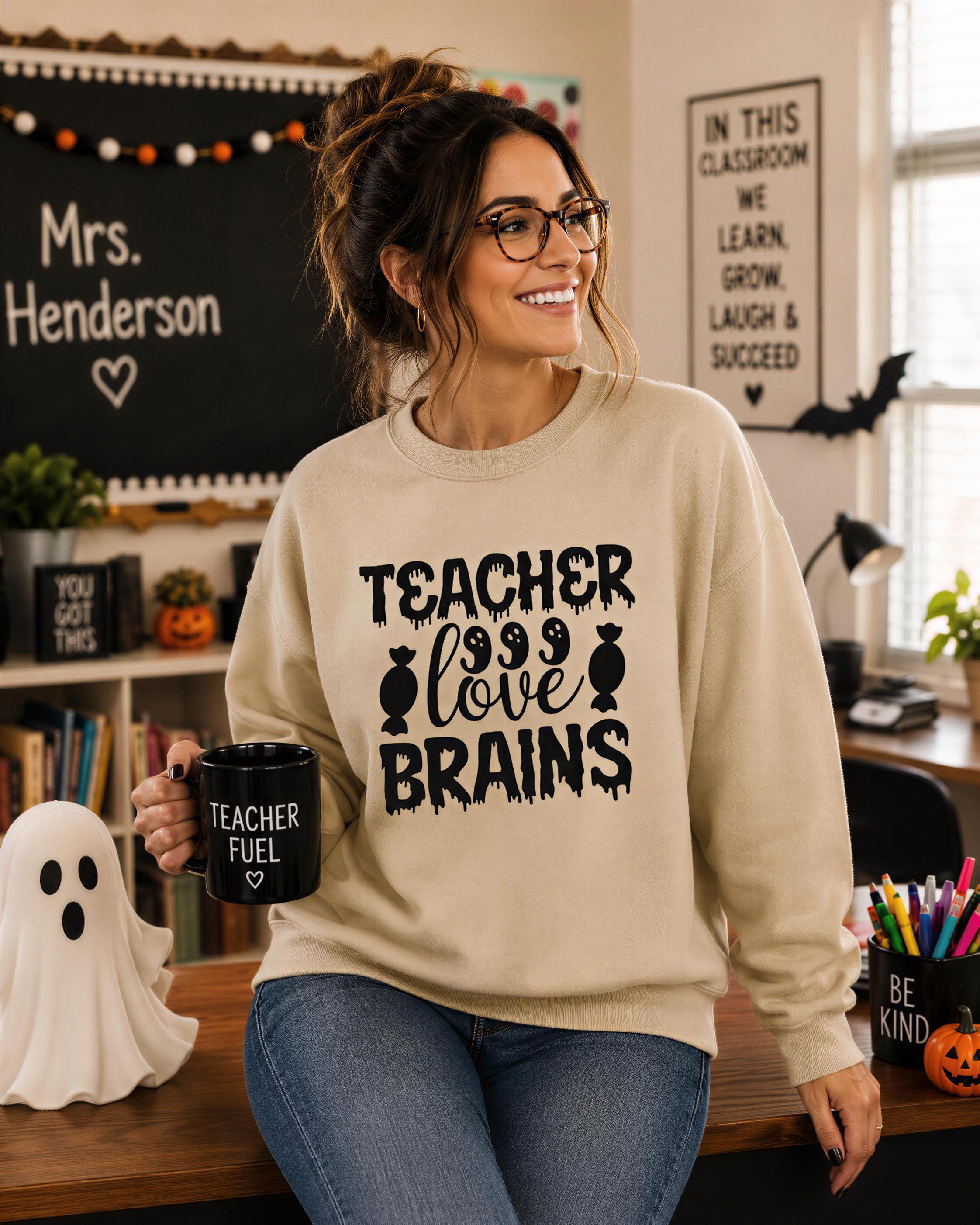 A woman wearing a beige sweater with the text "TEACHER LOVE BRAINS" is smiling and holding a mug with the same text. She is standing in a classroom setting with Halloween decorations, books, and a ghost.