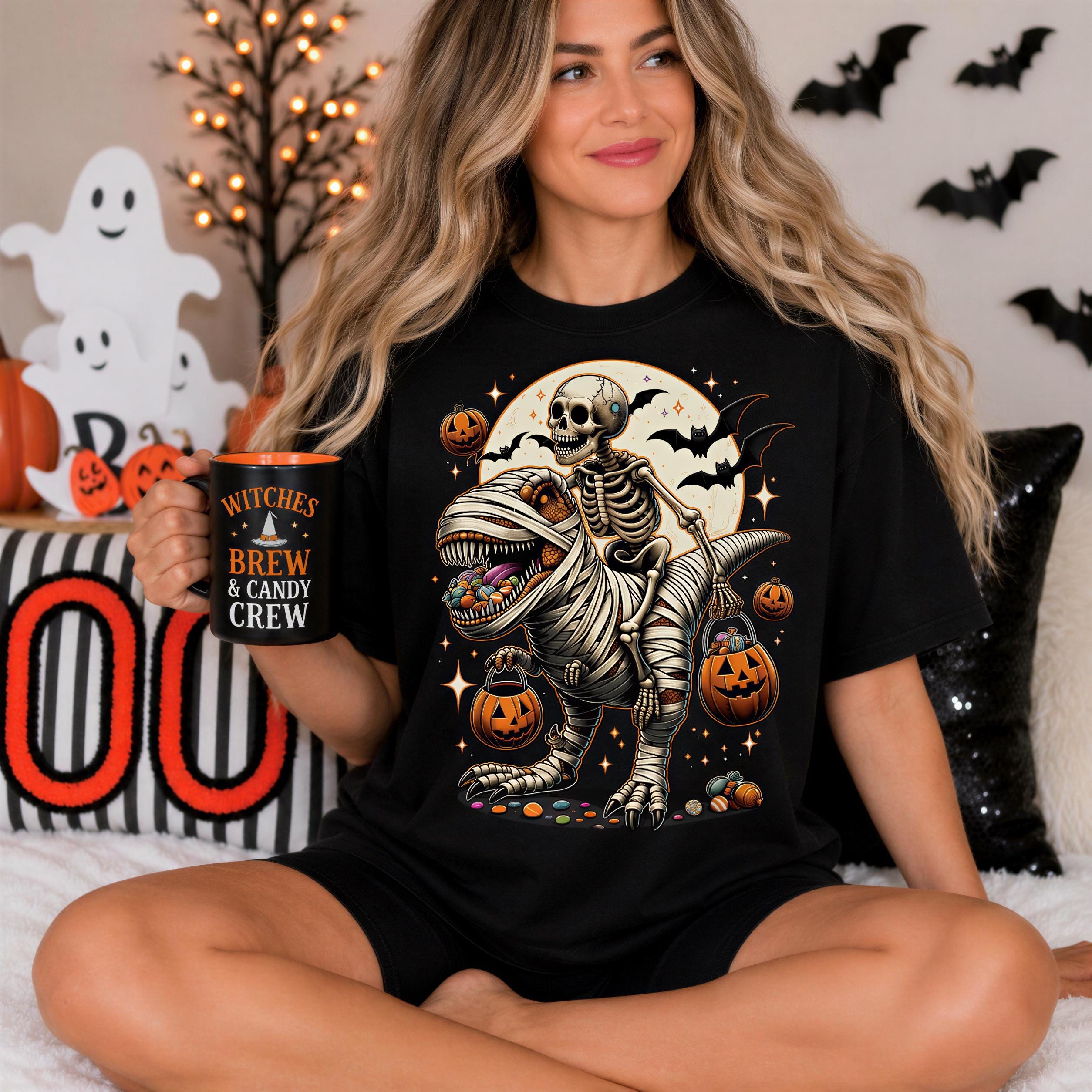 A woman is sitting on a bed, wearing a black t-shirt with a Halloween-themed graphic of a skeleton holding a skeleton-shaped pumpkin. She is holding a mug with the text "Witches Brew & Candy Crew" printed on it.