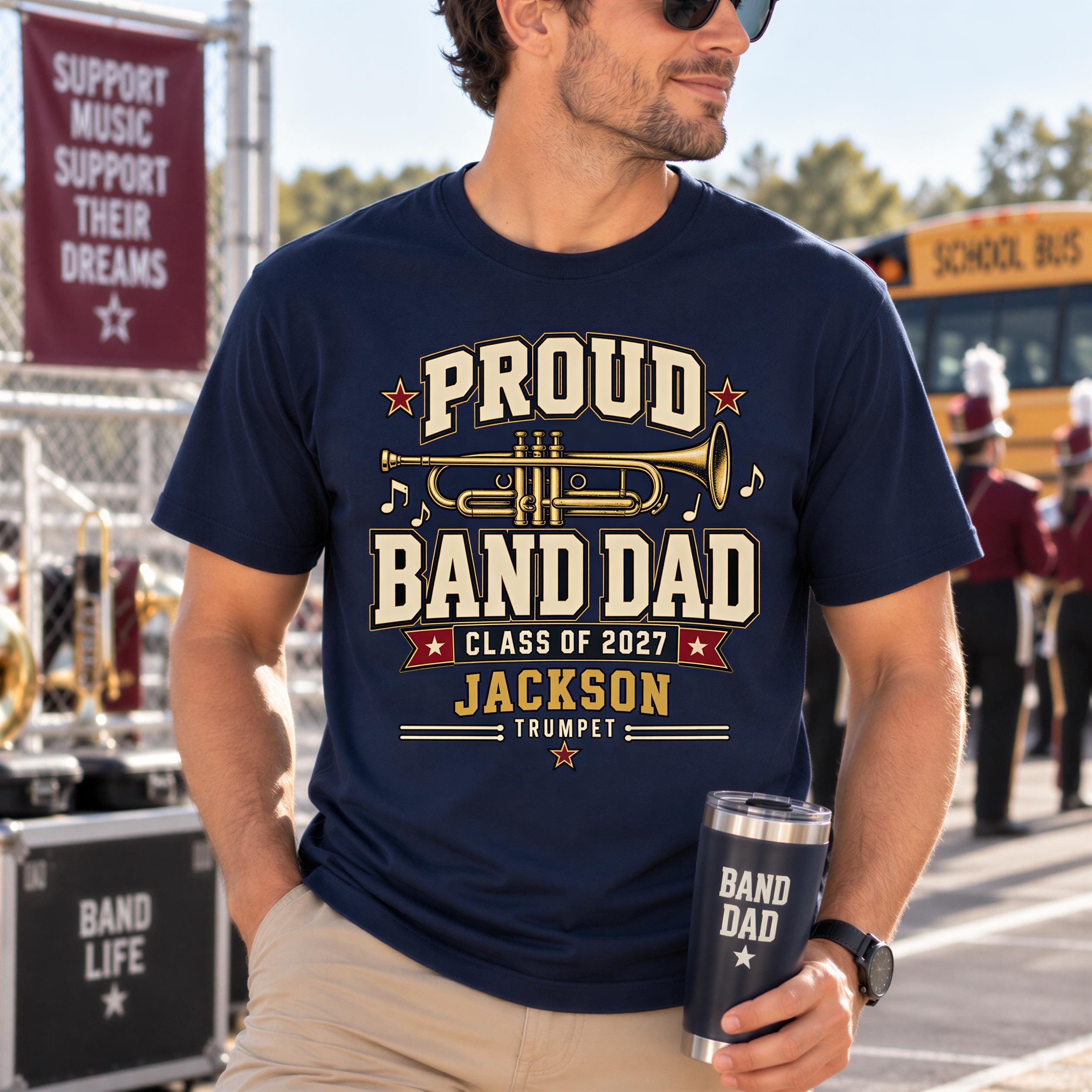 A man wearing a navy blue t-shirt with a graphic design that says "Proud Band Dad" and "Class of 2027 Jackson Trumpet" is holding a black tumbler with the text "BAND DAD" printed on it.