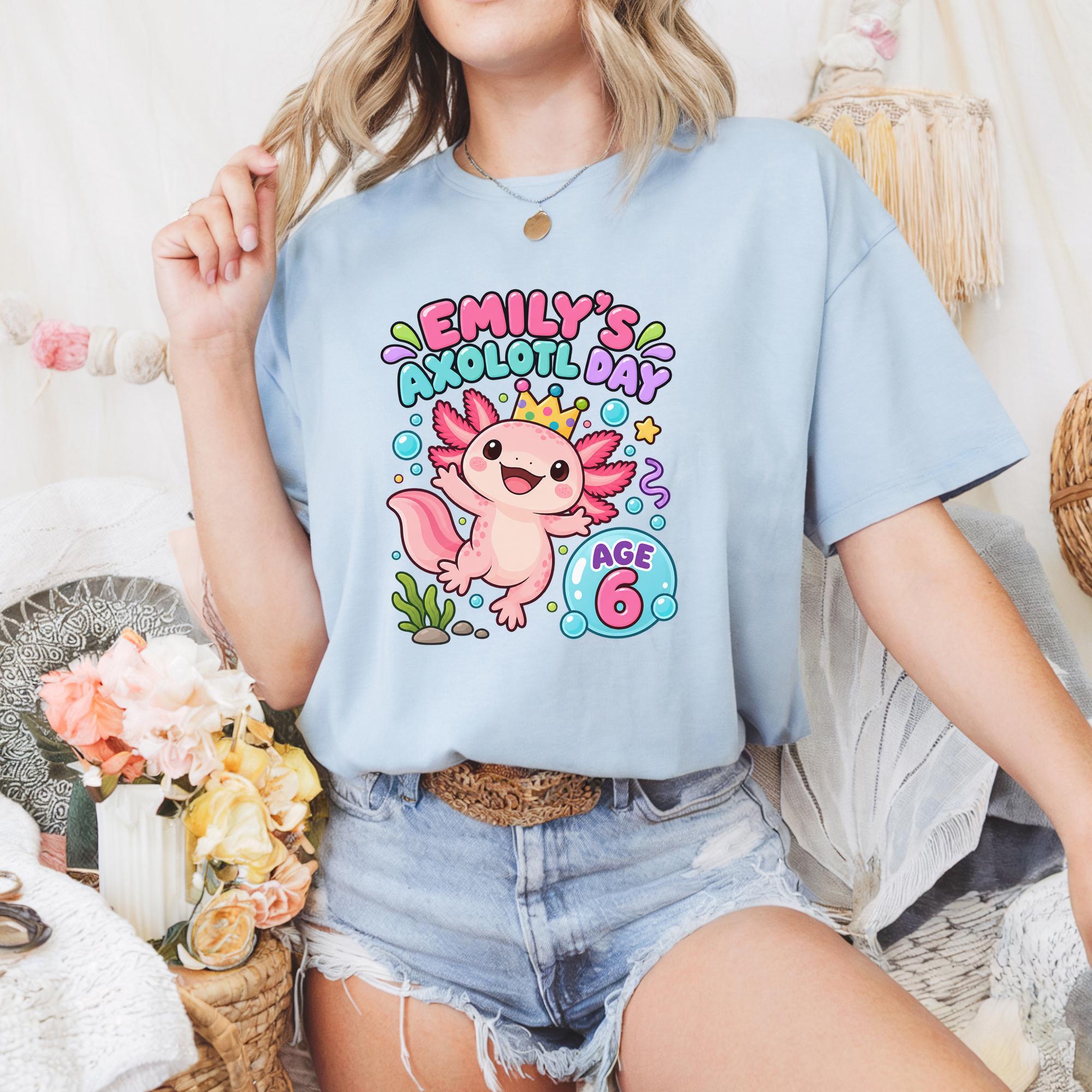 A woman wearing a light blue t-shirt with a colorful graphic of an axolotl, a type of salamander, and the text "Emilly's axolotl day age 6" printed on it.