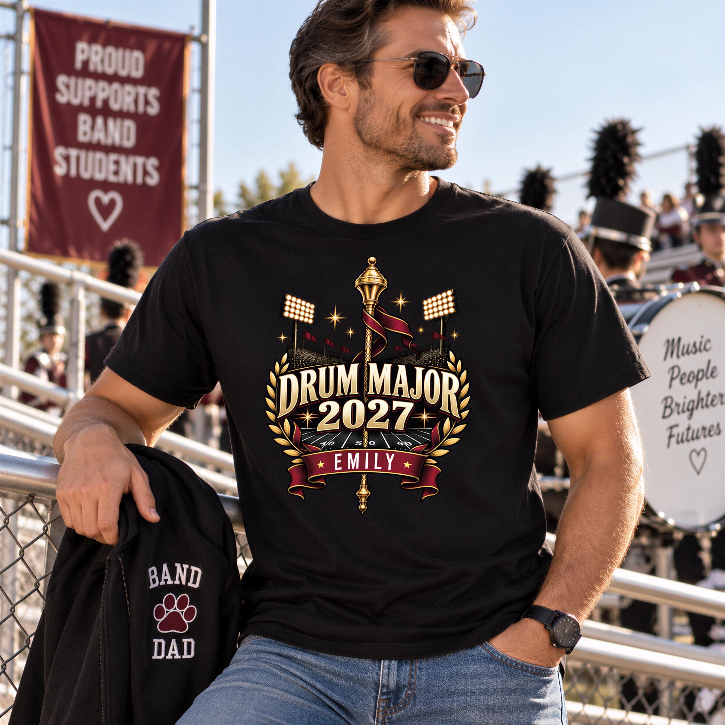 A man wearing a black t-shirt with a drum major design and a black jacket with a paw print and the text "BAND DAD" is standing in front of a crowd at what appears to be a music event.