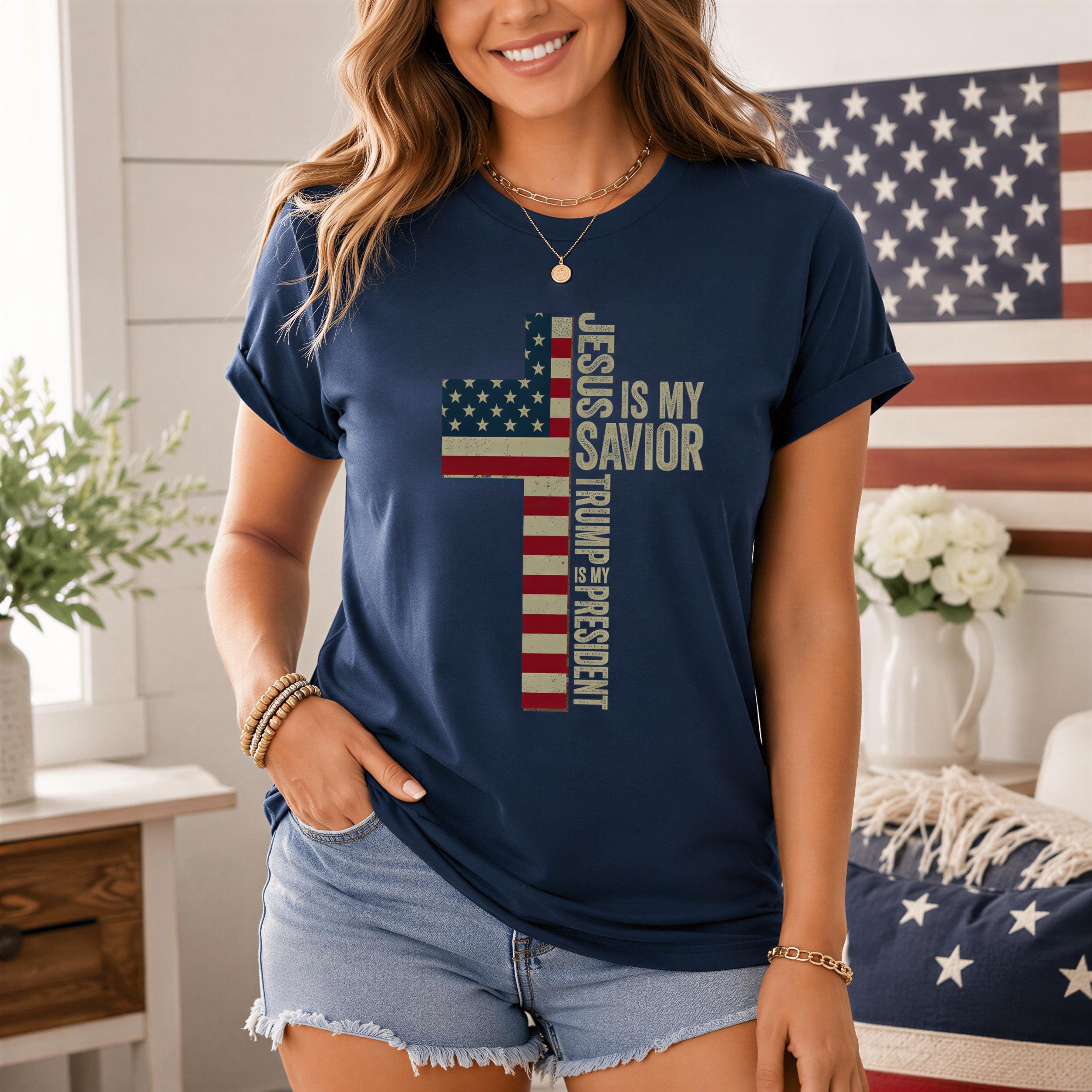 A woman wearing a navy blue t-shirt with a graphic design of a cross and the text "Jesus is my Savior Trump is president" stands in front of an American flag.