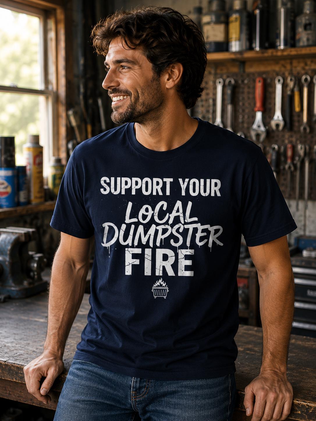 A man wearing a navy blue t-shirt with the text "Support Your Local Dumpster Fire" is smiling and leaning against a workbench in a workshop setting.