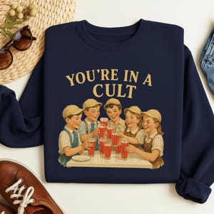 A navy blue sweatshirt with a graphic design depicting a group of children happily drinking from glasses, accompanied by the text "YOU'RE IN A CULT".