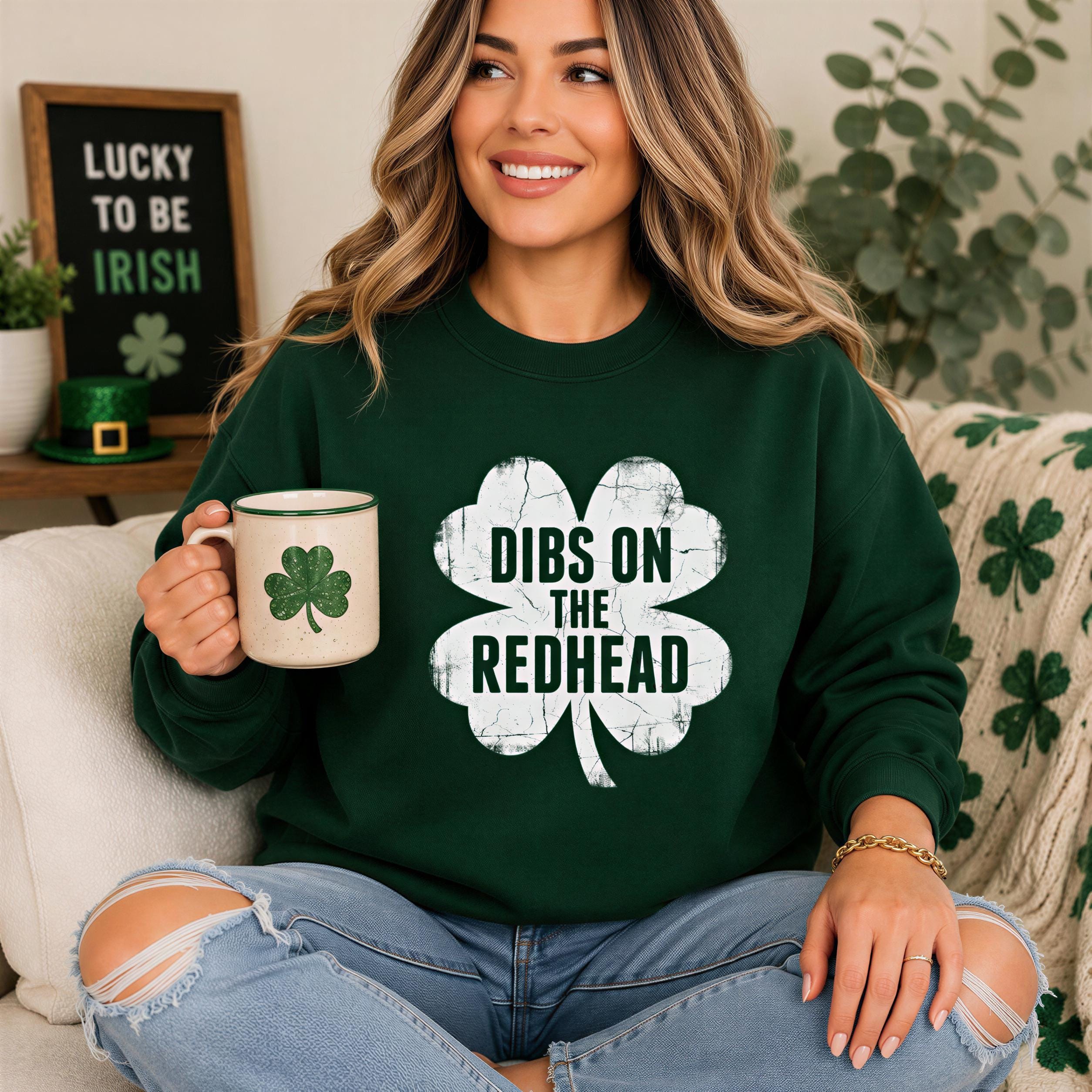 A woman wearing a green sweatshirt with the text "DIBS ON THE REDHEAD" is sitting on a couch, holding a mug with a clover design.