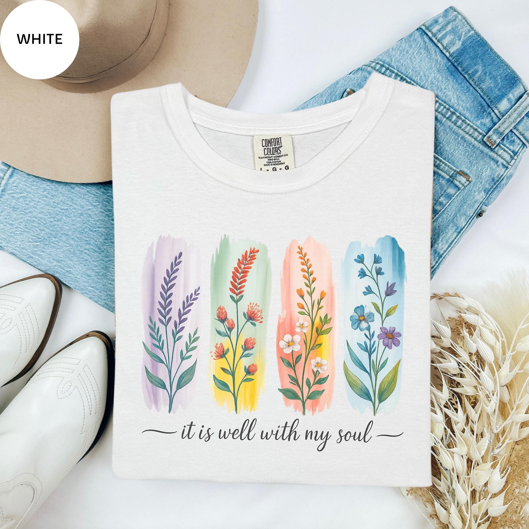 a white t-shirt with a colorful graphic design of four different flowers, accompanied by the text "it is well with my soul". The t-shirt is laid out on a white surface, with a pair of jeans, a hat, and some flowers nearby.