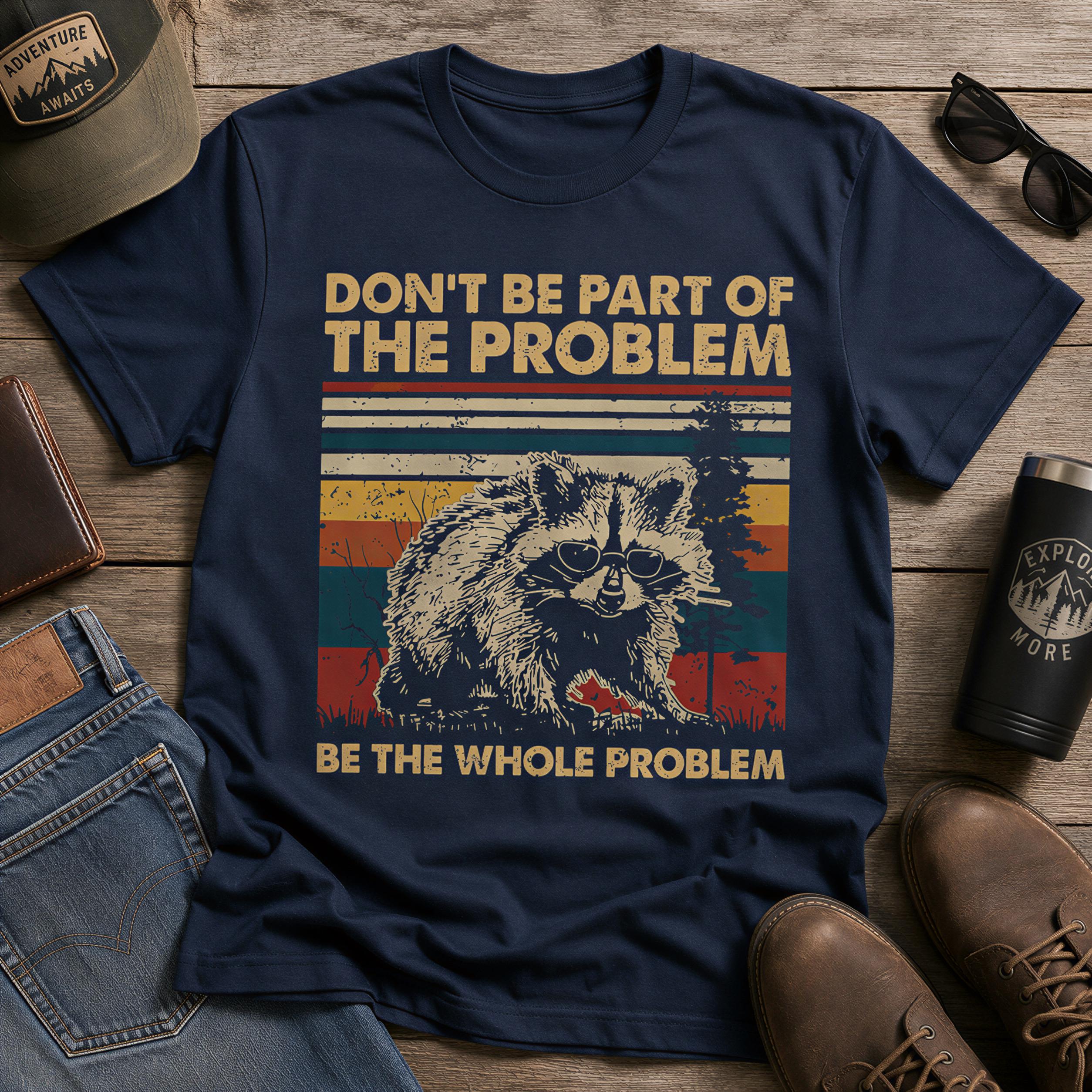 A navy blue t-shirt with a graphic design of a raccoon and the text "DON'T BE PART OF THE PROBLEM BE THE WHOLE PROBLEM" printed on it, along with a vintage-style graphic. The t-shirt is laid out on a wooden surface, accompanied by a pair of jeans, a hat, sunglasses, and a pair of shoes.