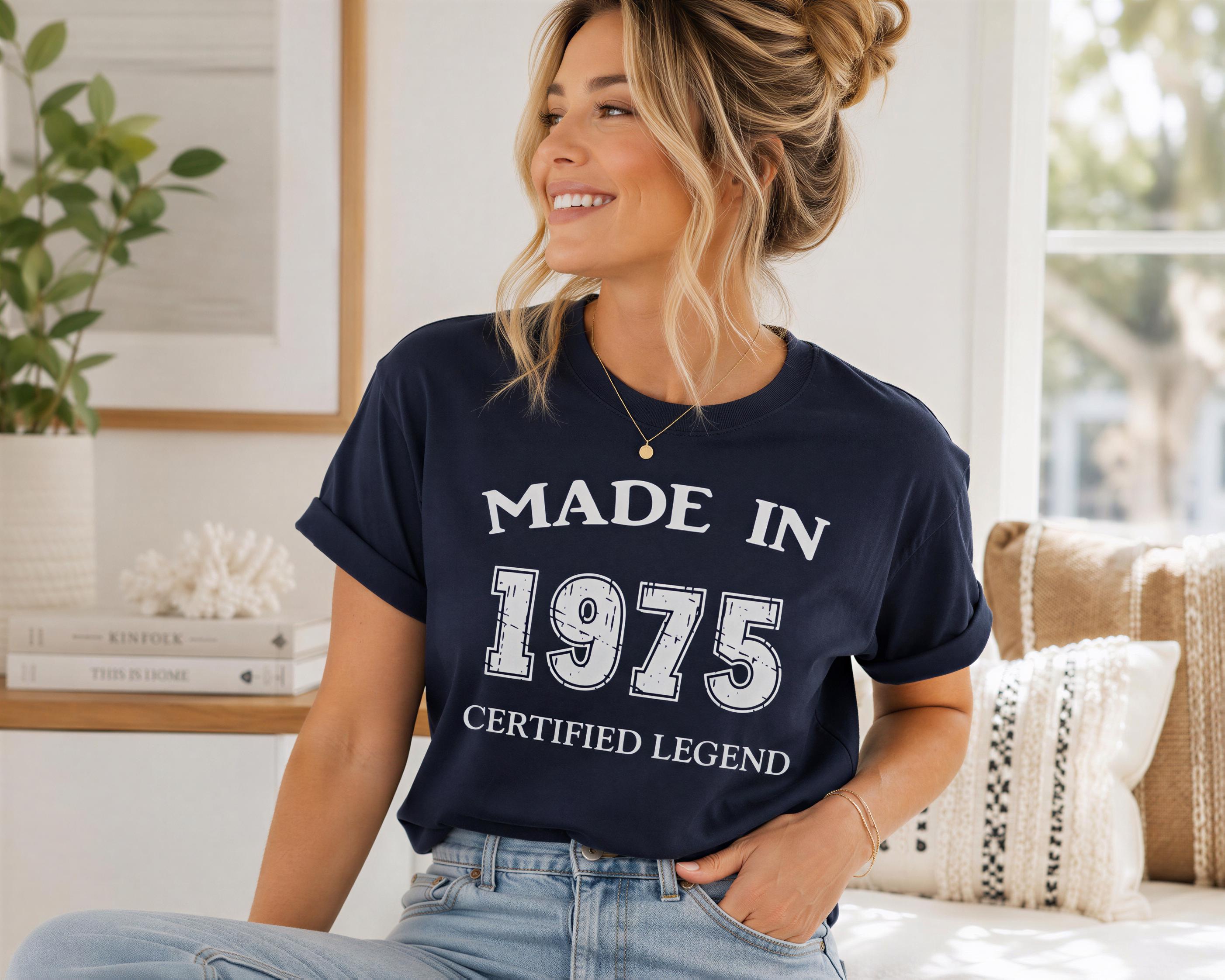 A smiling woman wearing a navy blue t-shirt with the text "MADE IN 1975 CERTIFIED LEGEND" is sitting on a couch in a room with a potted plant and books.
