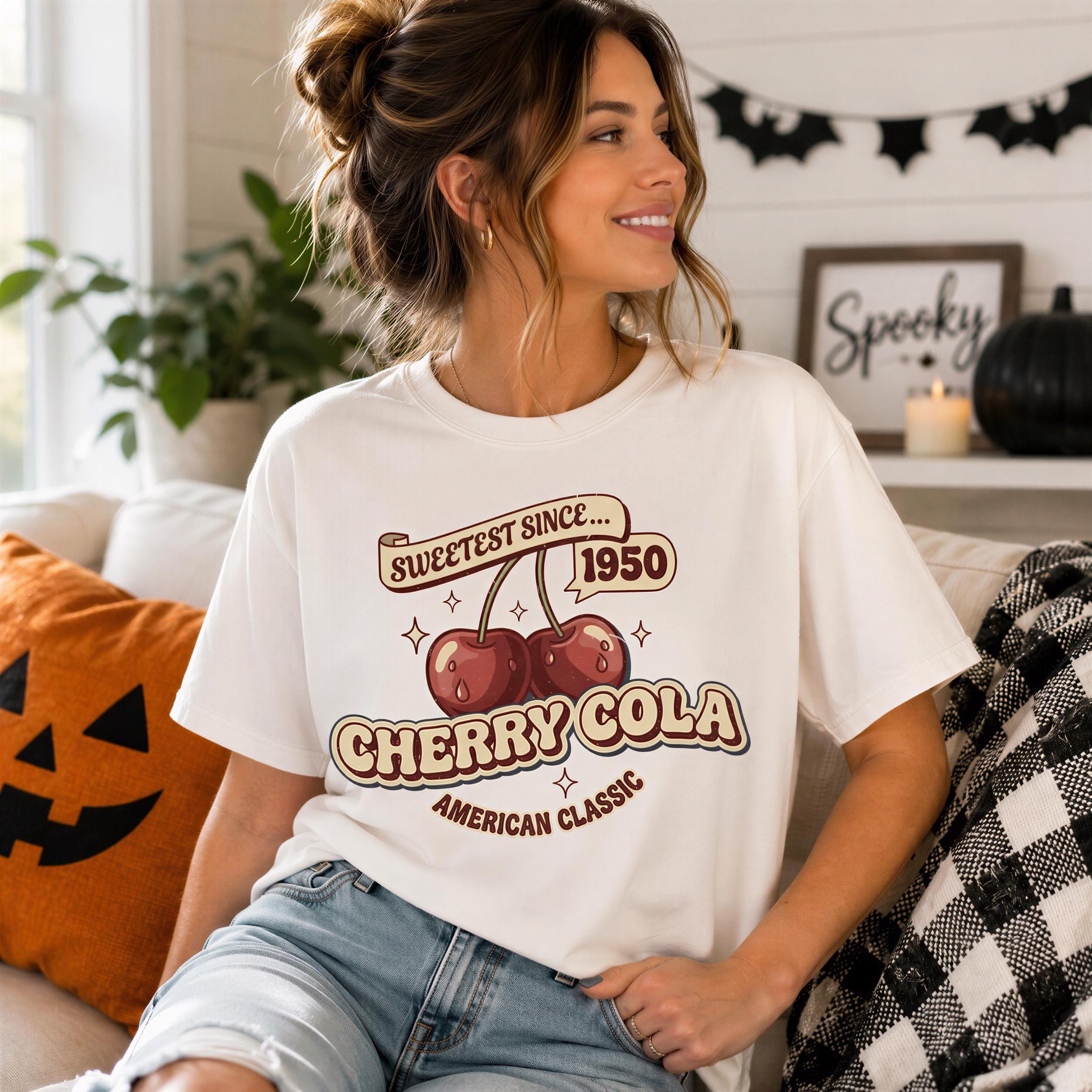A woman wearing a white t-shirt with a graphic design of cherries and a text that says "Sweetest since 1950 Cherry Cola American Classic" is sitting on a couch.