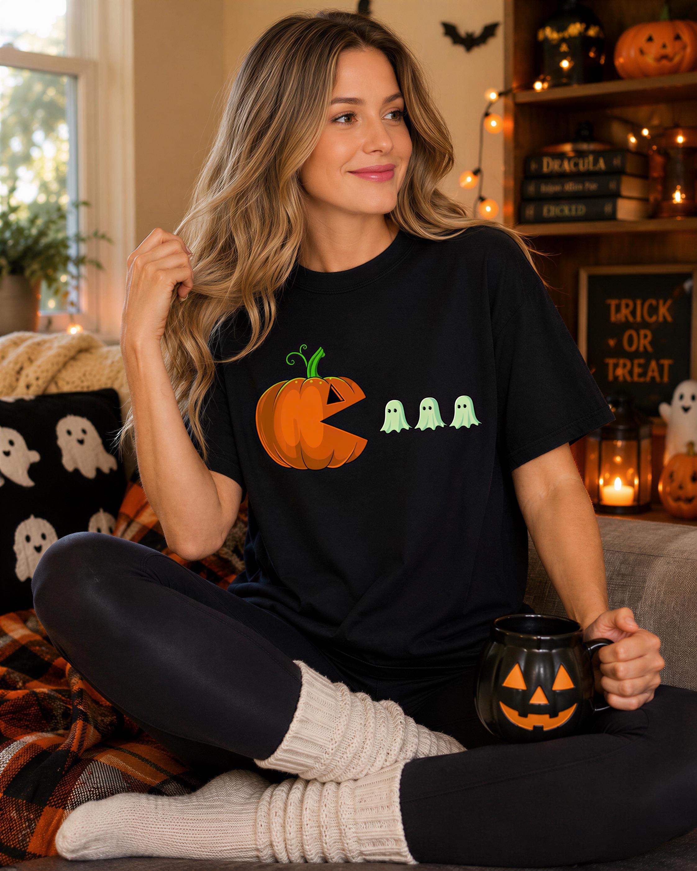 A woman wearing a black t-shirt with a pumpkin graphic and ghost figures is sitting on a couch, holding a mug with a jack-o-lantern design.
