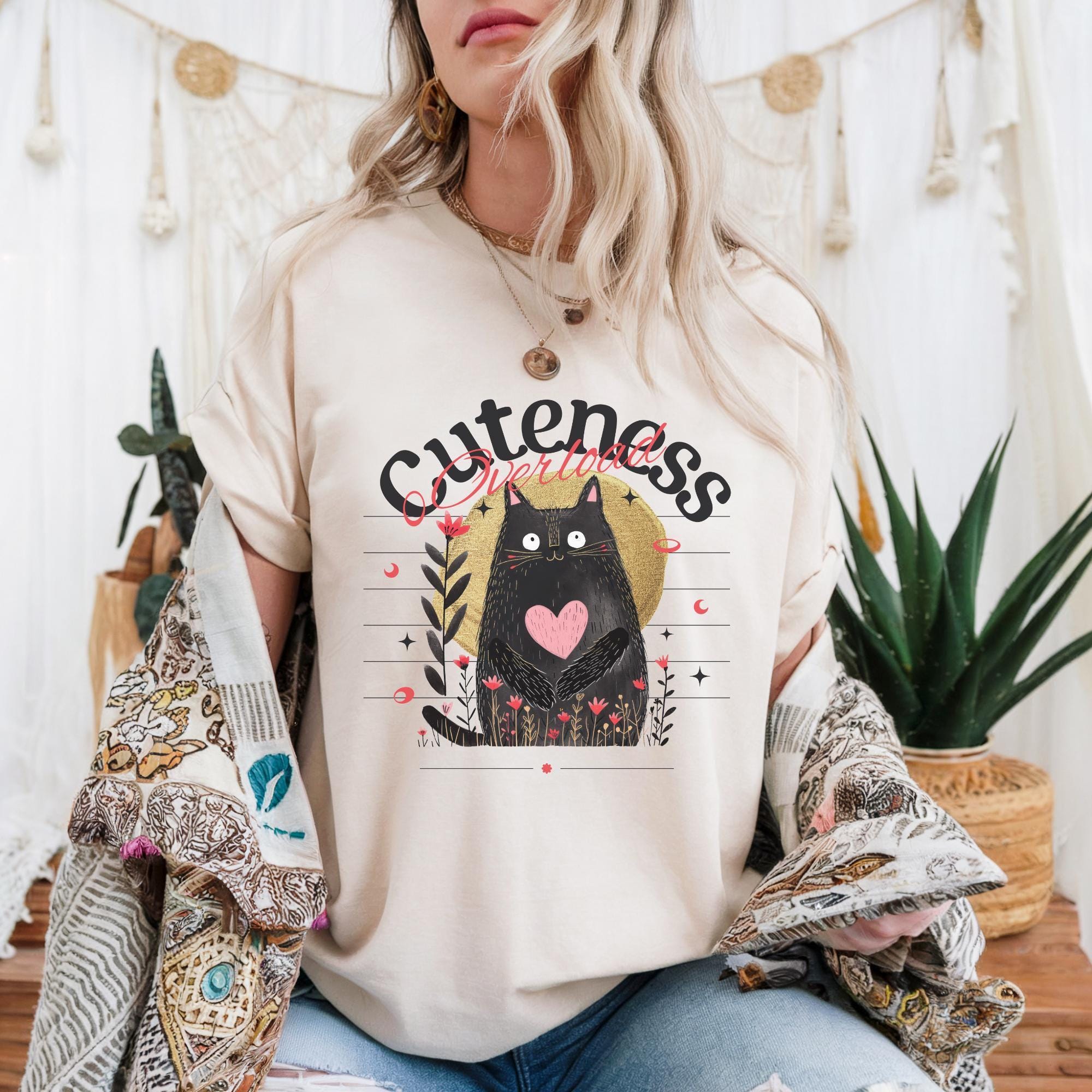 A woman wearing a white t-shirt with a graphic design of a black cat and the text "Cuteness" is sitting in front of a white curtain with plants and a string of lights.