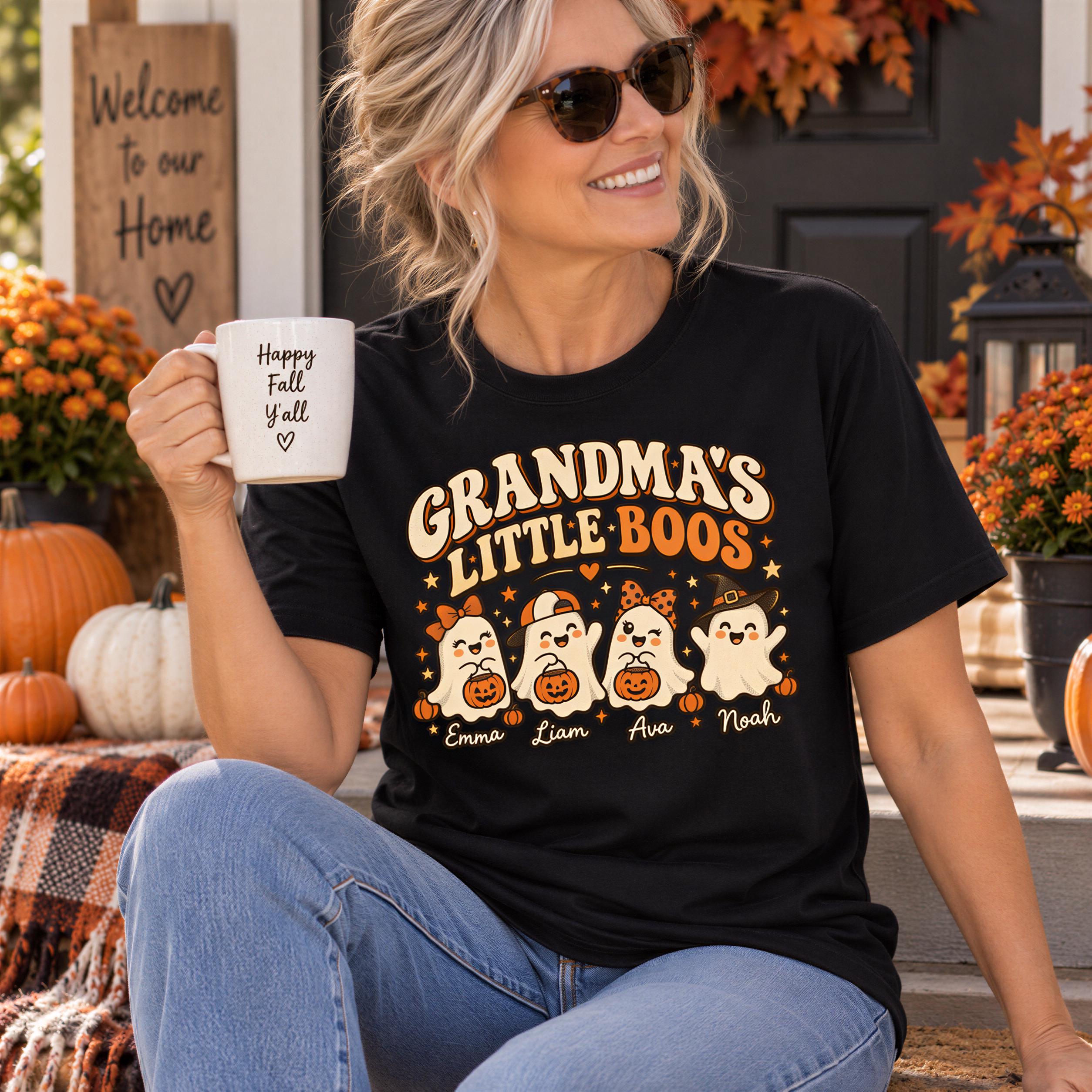 A woman wearing a black t-shirt with a graphic of a group of ghostly figures and the text "Grandma's Little Boos" is sitting on a porch surrounded by fall decorations. She is holding a coffee mug with the text "Happy Fall Yell" printed on it.