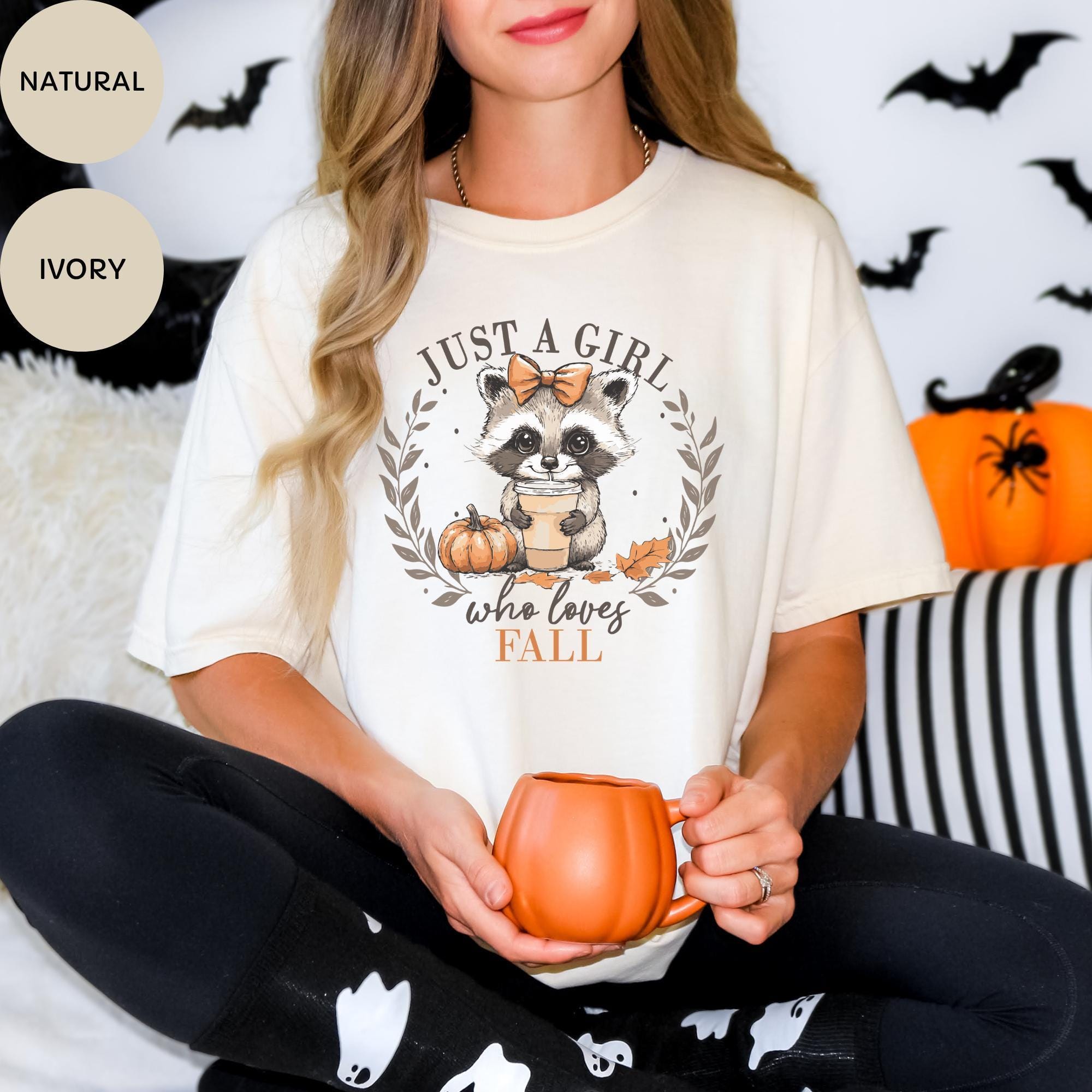 A woman wearing a white t-shirt with a graphic of a raccoon holding a pumpkin and the text "Just a girl who loves fall" is sitting on a couch with a mug in her hand.