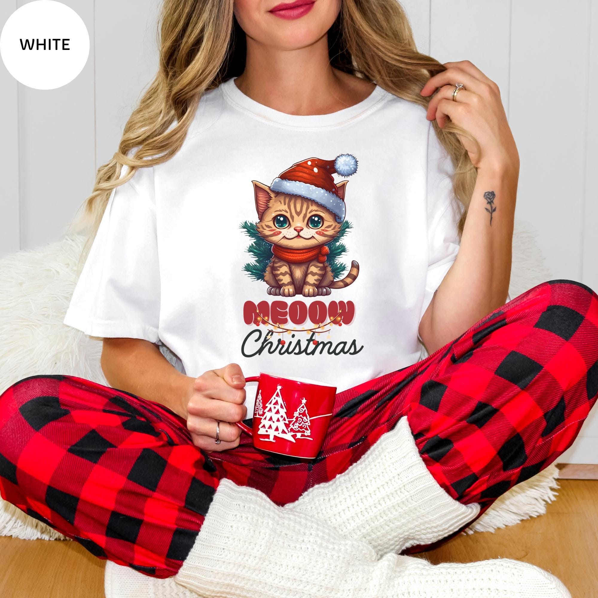 A woman wearing a white t-shirt with a cat graphic and the text "Meow on Christmas" is sitting on the floor, holding a mug.
