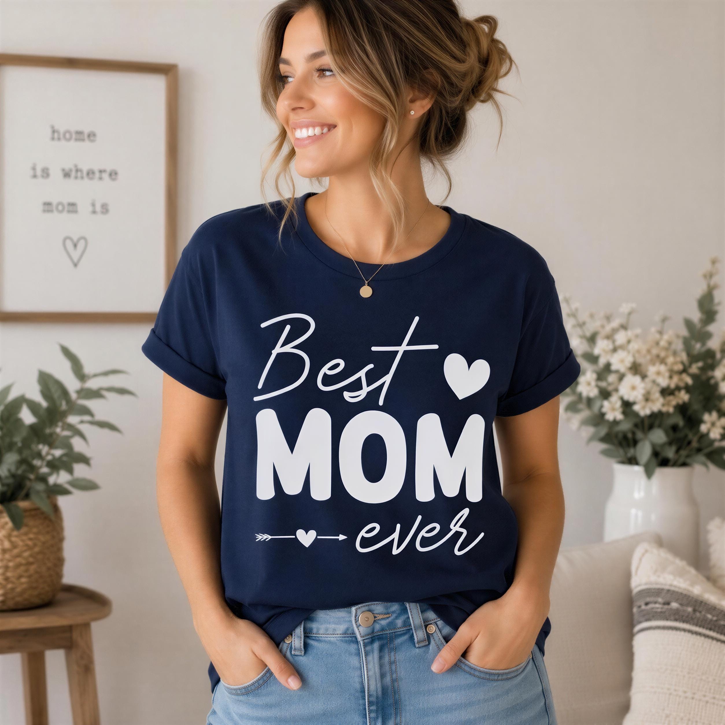 A woman wearing a navy blue t-shirt with the text "Best Mom Ever" is smiling and posing in a room with a framed quote and flowers.
