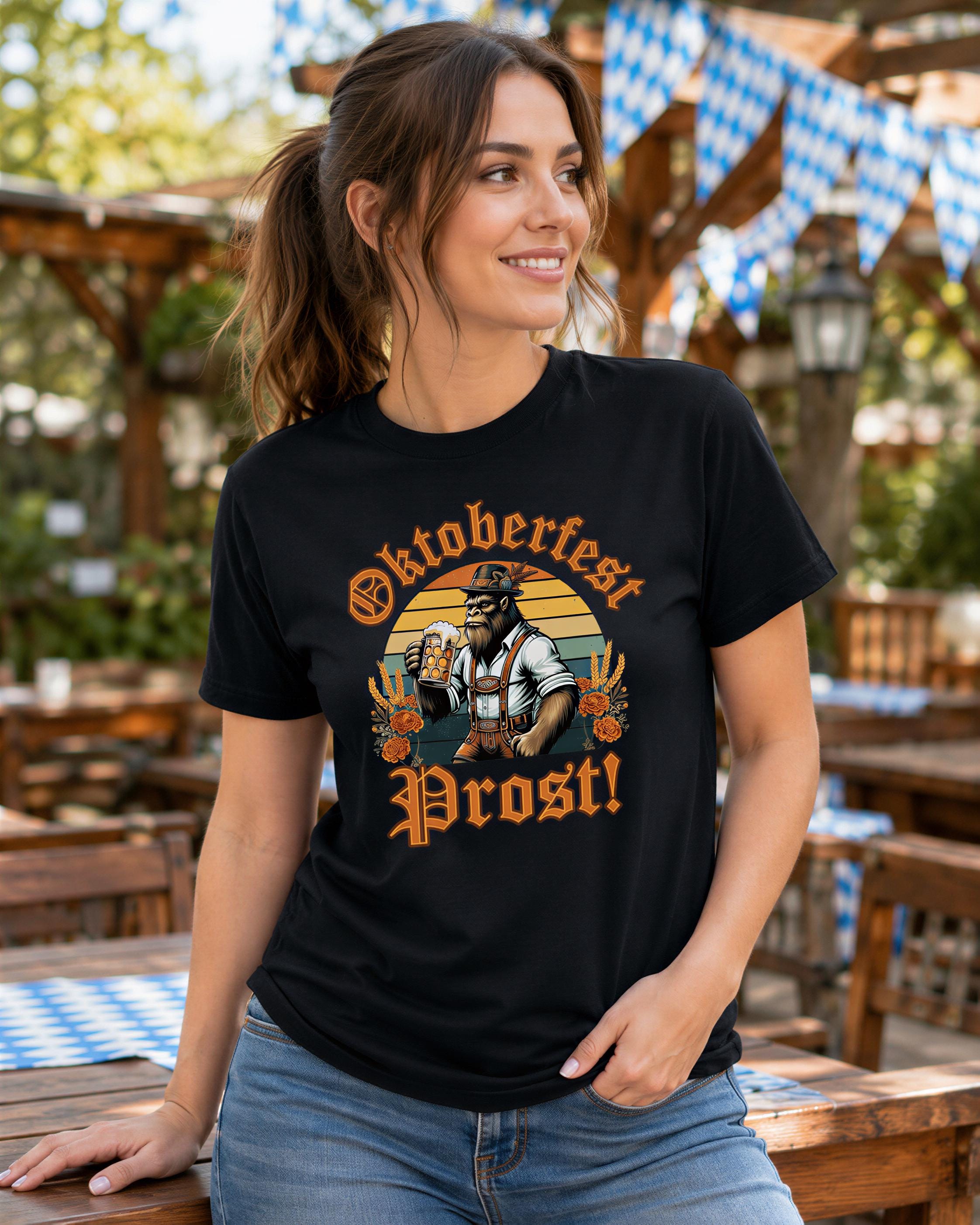 A woman wearing a black t-shirt with an illustration of a man holding a beer and the text "Oktoberfest Prost!" is smiling and posing in an outdoor setting with tables and chairs.
