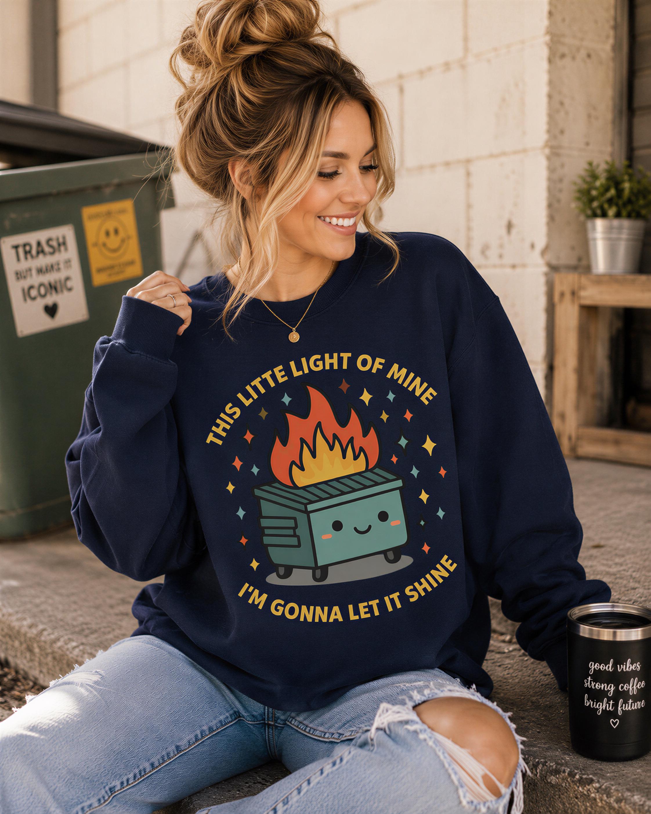 A woman wearing a navy blue sweatshirt with a graphic design of a fire-breathing trash can and the text "This little light of mine" is sitting on the ground.