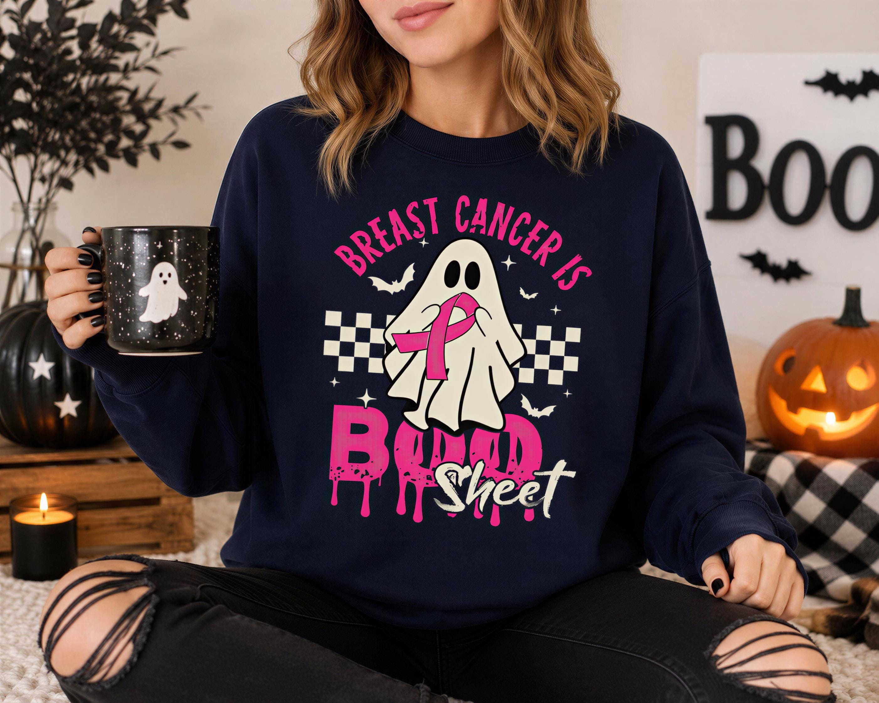 A woman wearing a black sweatshirt with a graphic design that says "BREAST CANCER IS BIND SHEET" and holding a mug with a ghost design.