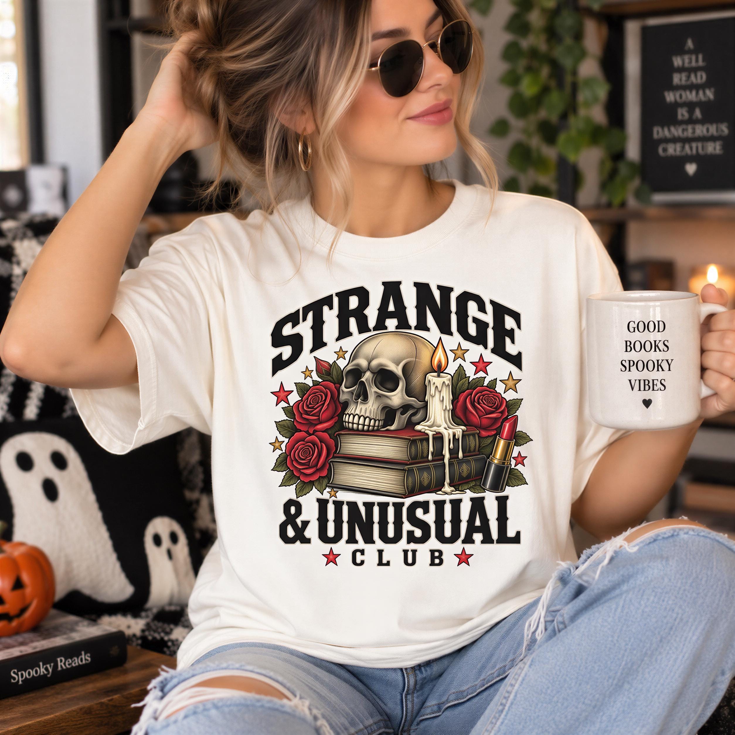 A woman wearing a white t-shirt with a graphic design and holding a mug with text on it, sitting in a room with Halloween decorations and books.