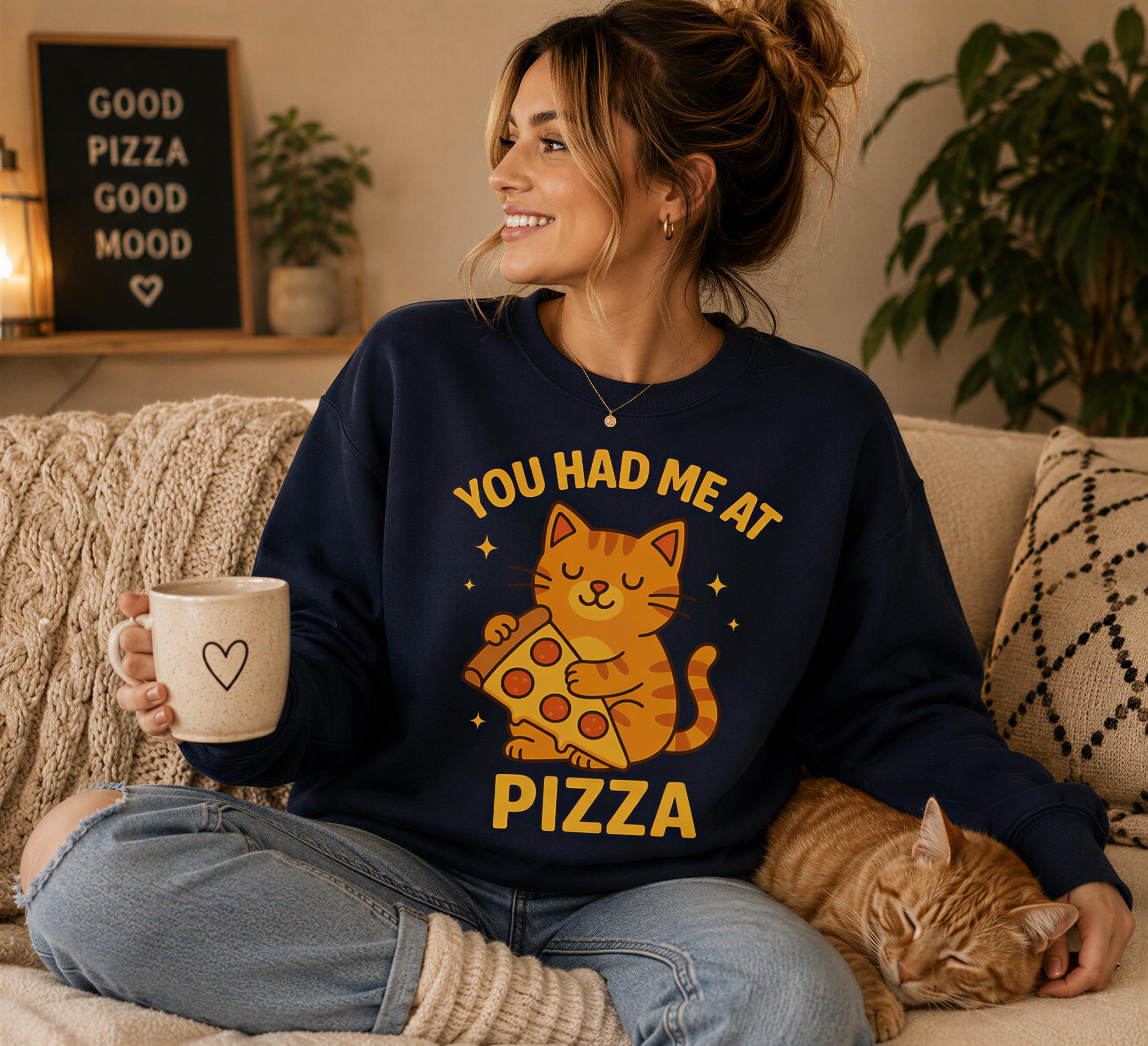 A woman sitting on a couch, wearing a sweatshirt with a cat graphic and the text "YOU HAD ME AT PIZZA". She is holding a coffee mug and has a cat sleeping on her lap.