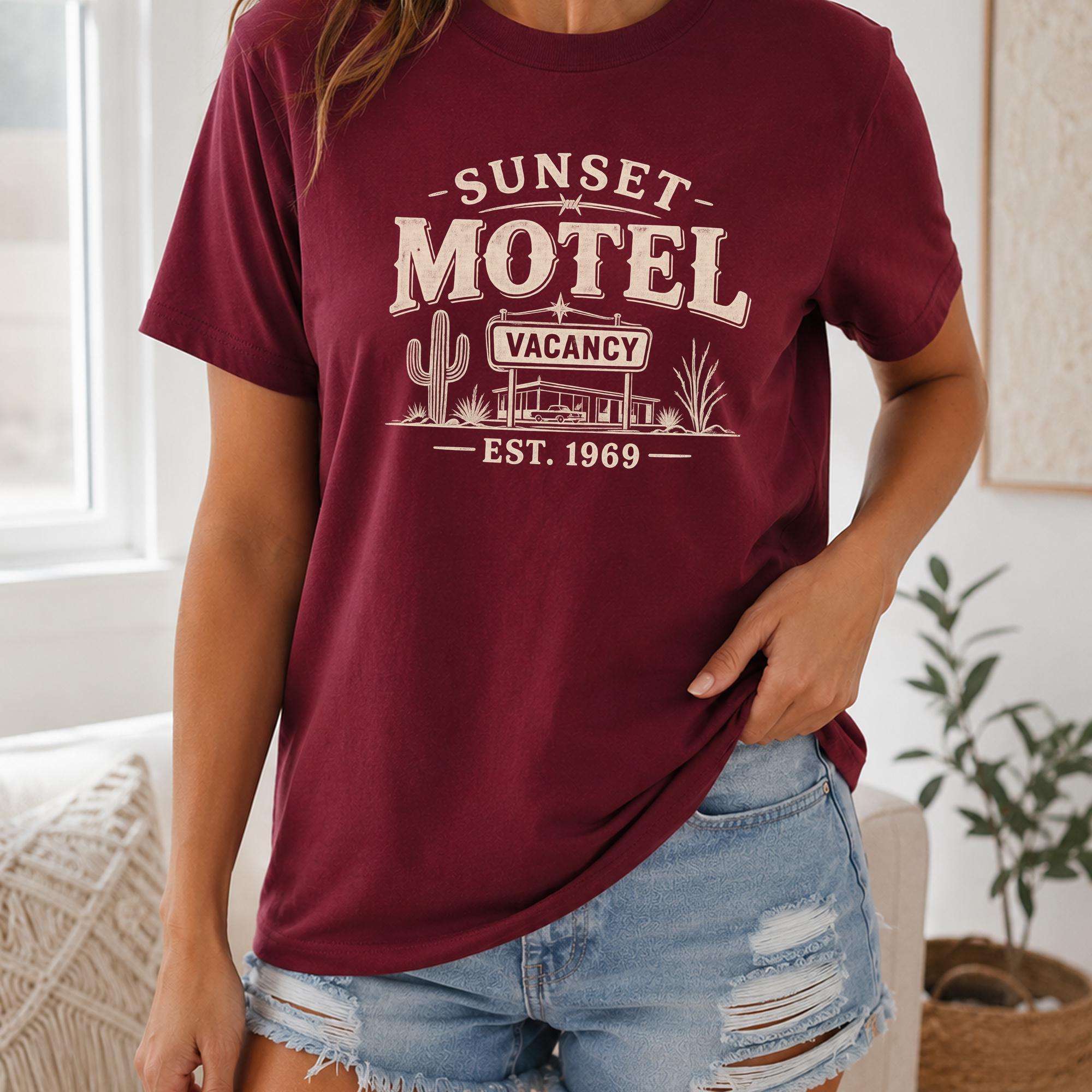 A woman wearing a burgundy t-shirt with a graphic design of a motel and a cactus, standing in front of a window.