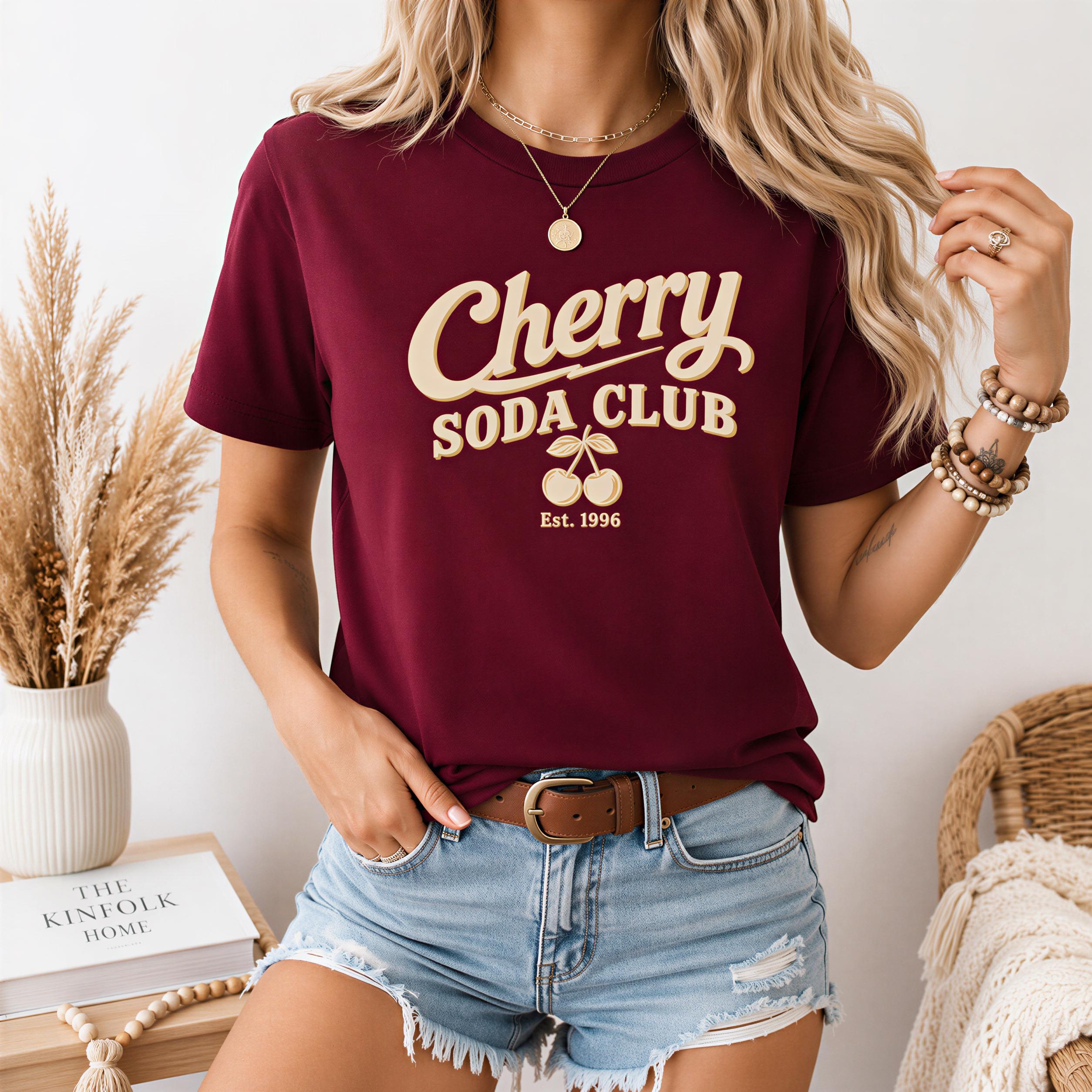 A woman wearing a burgundy t-shirt with the text "Cherry Soda Club" and denim shorts, standing in front of a white wall with a vase of dried flowers and a wooden table.