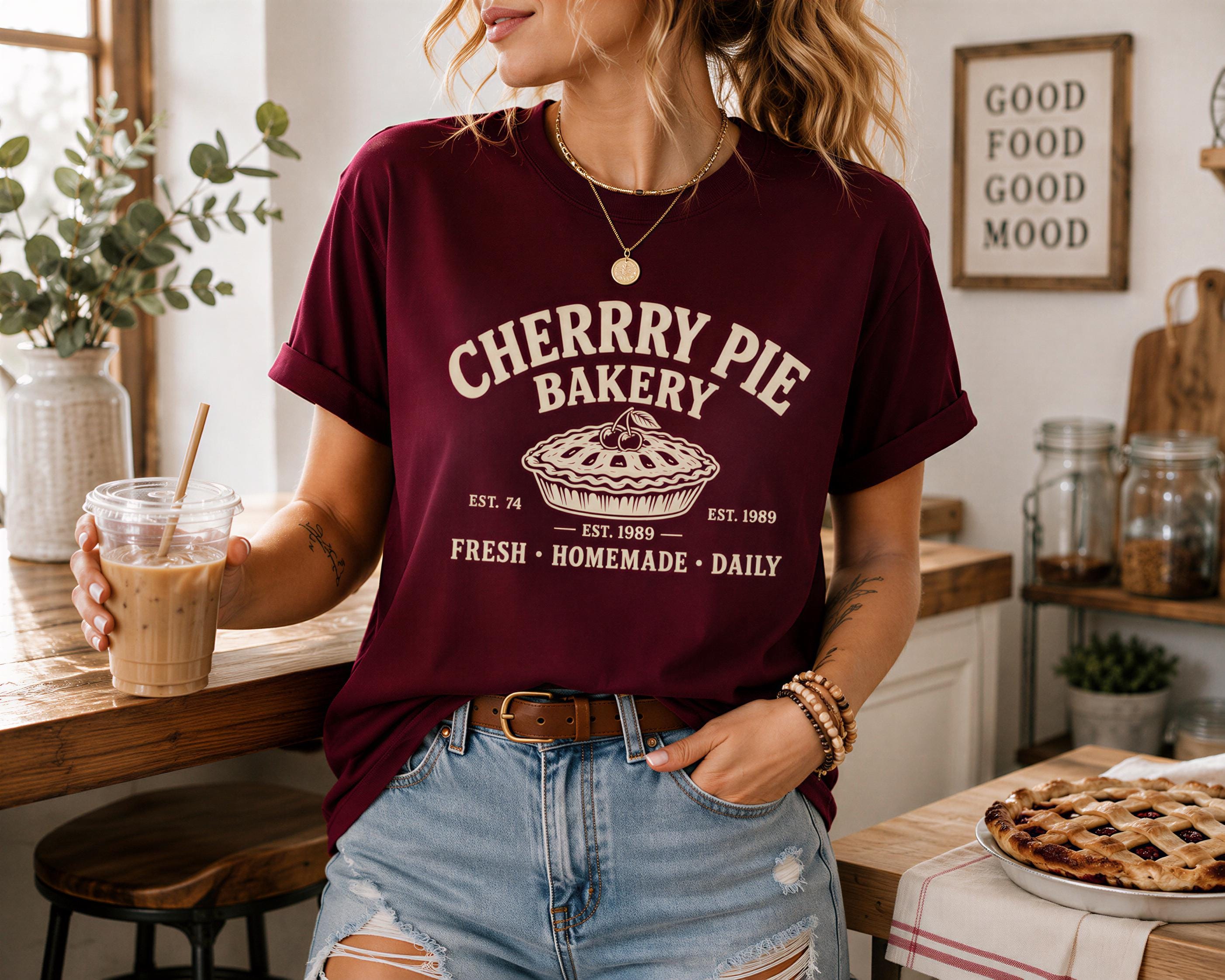 A woman wearing a burgundy t-shirt with the text "CHERRY PIE BAKERY" and holding a drink stands in a kitchen with a pie on the counter.