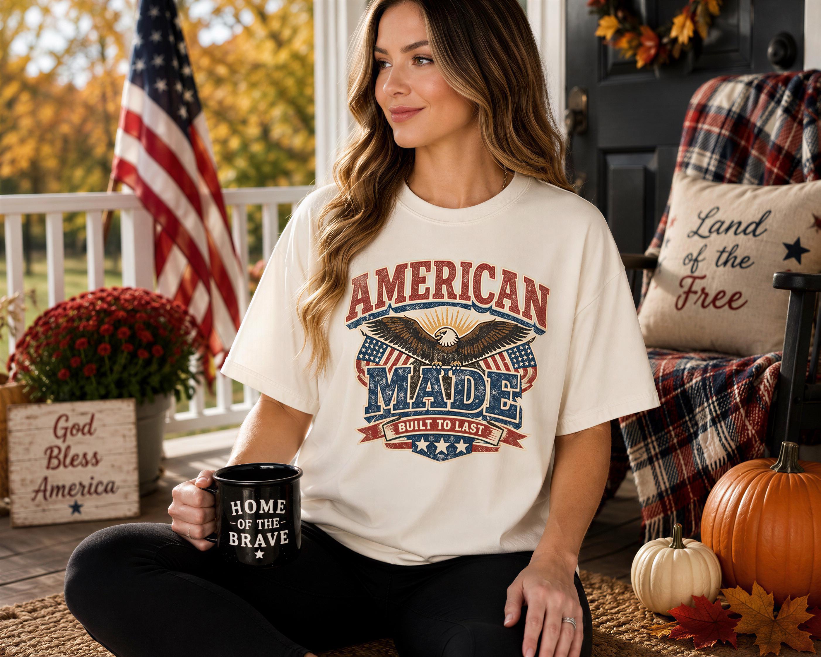 A woman wearing a white t-shirt with an American flag design and the text "AMERICAN MADE" is sitting on a porch, holding a black mug with the text "HOME OF THE BRAVE". The porch is decorated with American flags, pumpkins, and other fall-themed items, creating a patriotic and cozy atmosphere.