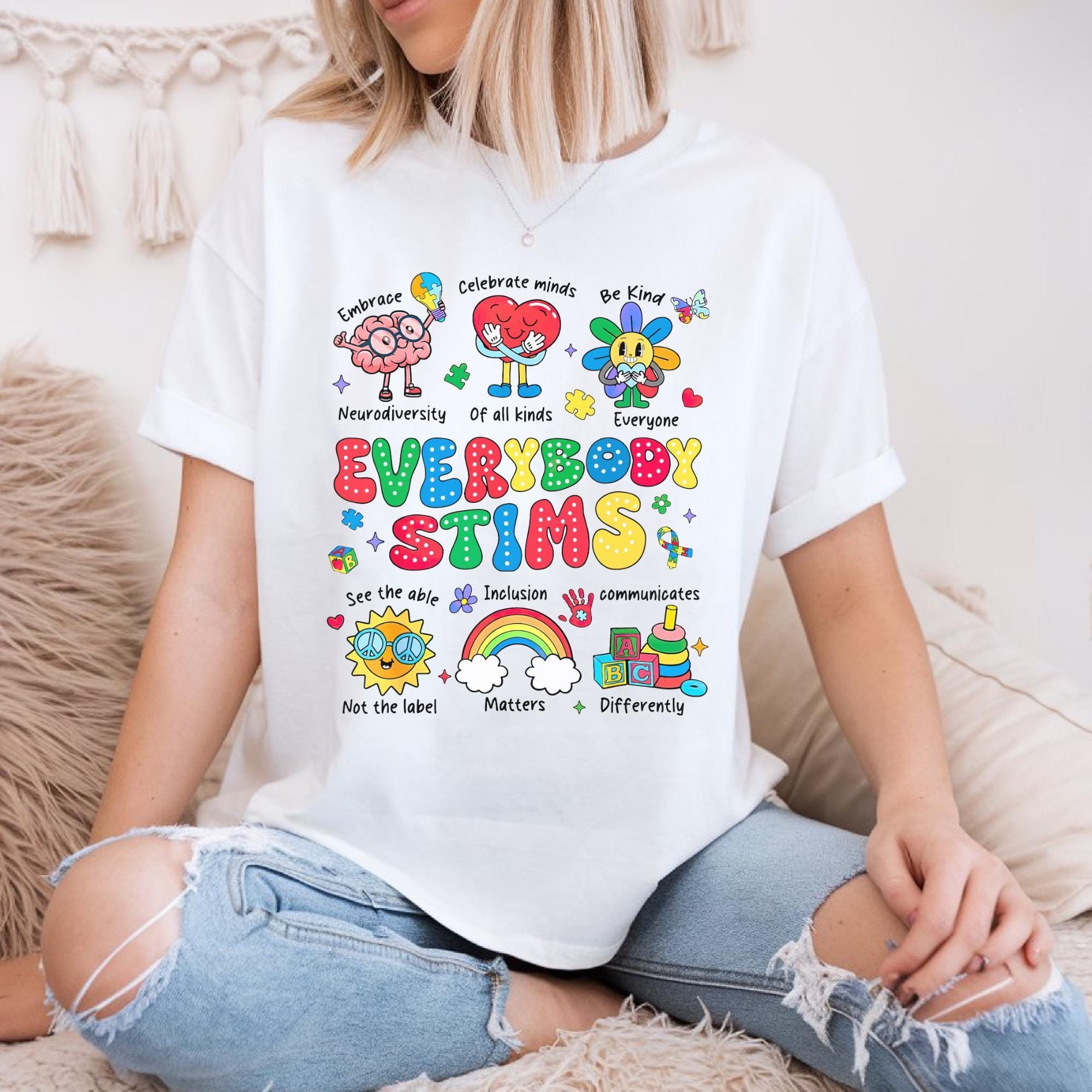 A person wearing a white t-shirt with a colorful graphic design featuring various characters and text related to the concept of "Everybody Stims".