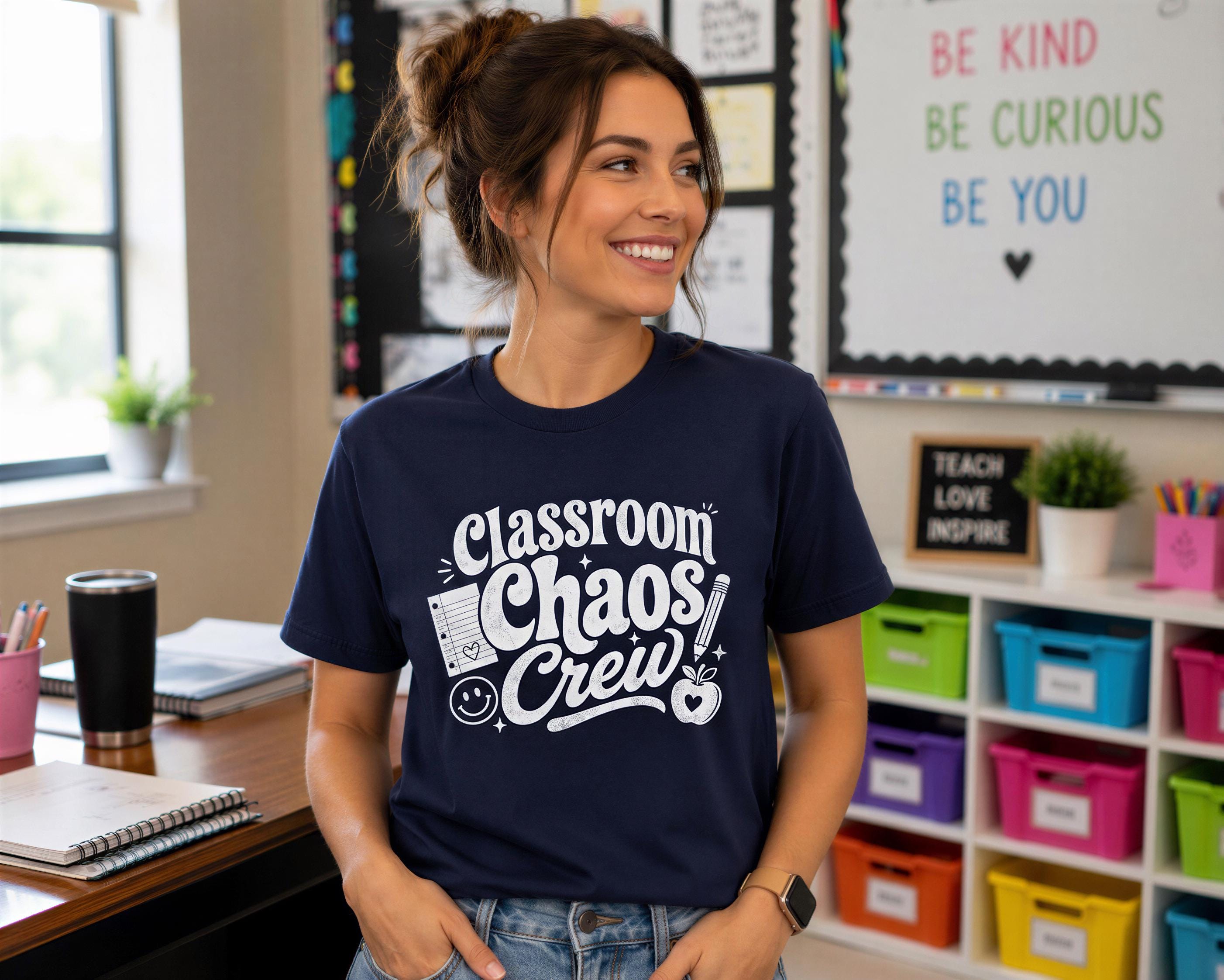 A woman wearing a navy blue t-shirt with the text "Classroom Chaos Crew" stands in a classroom with a bulletin board and colorful bins in the background.
