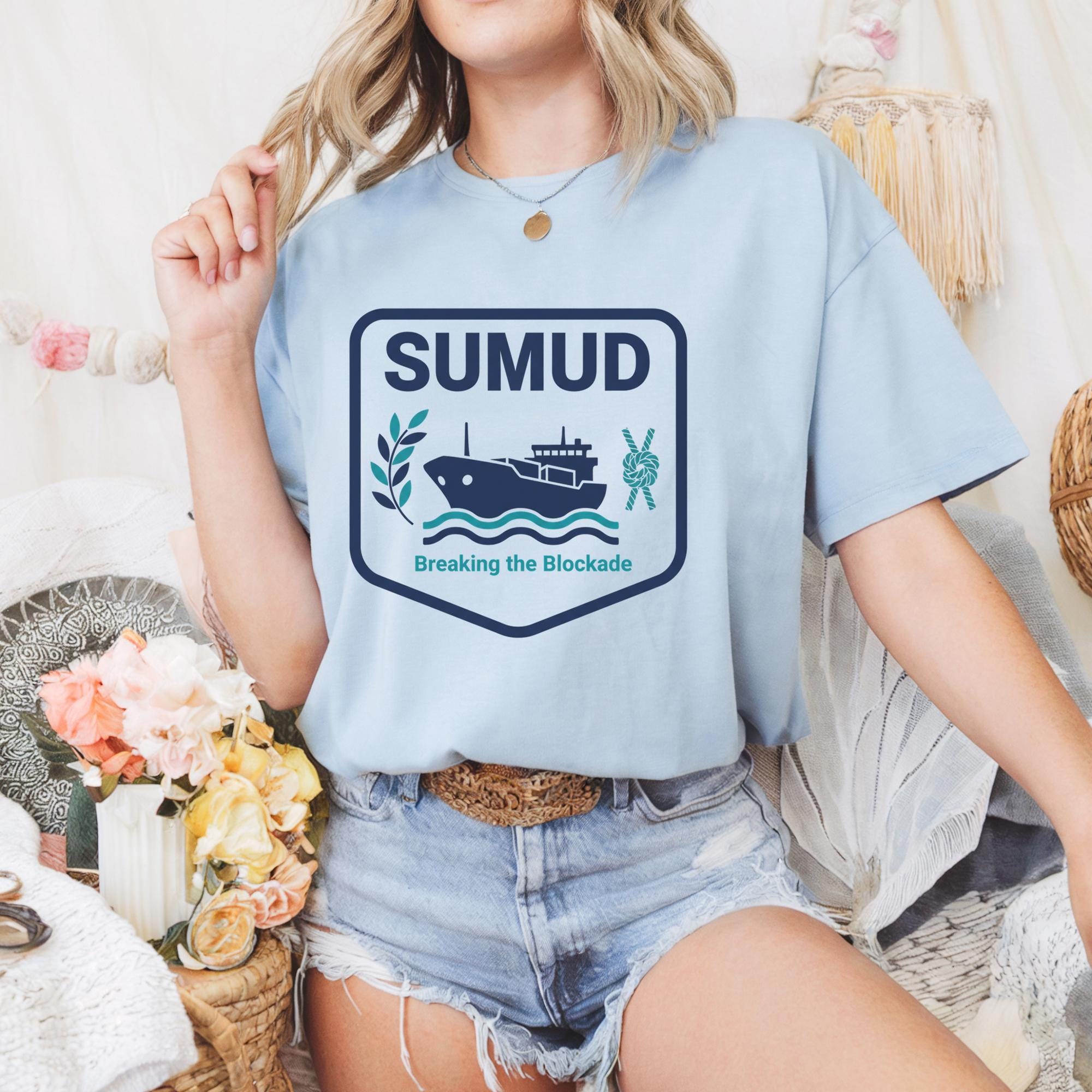 A woman wearing a light blue t-shirt with a graphic design of a ship and the text "SUMUD Breaking the Blockade" is sitting on a chair surrounded by flowers.