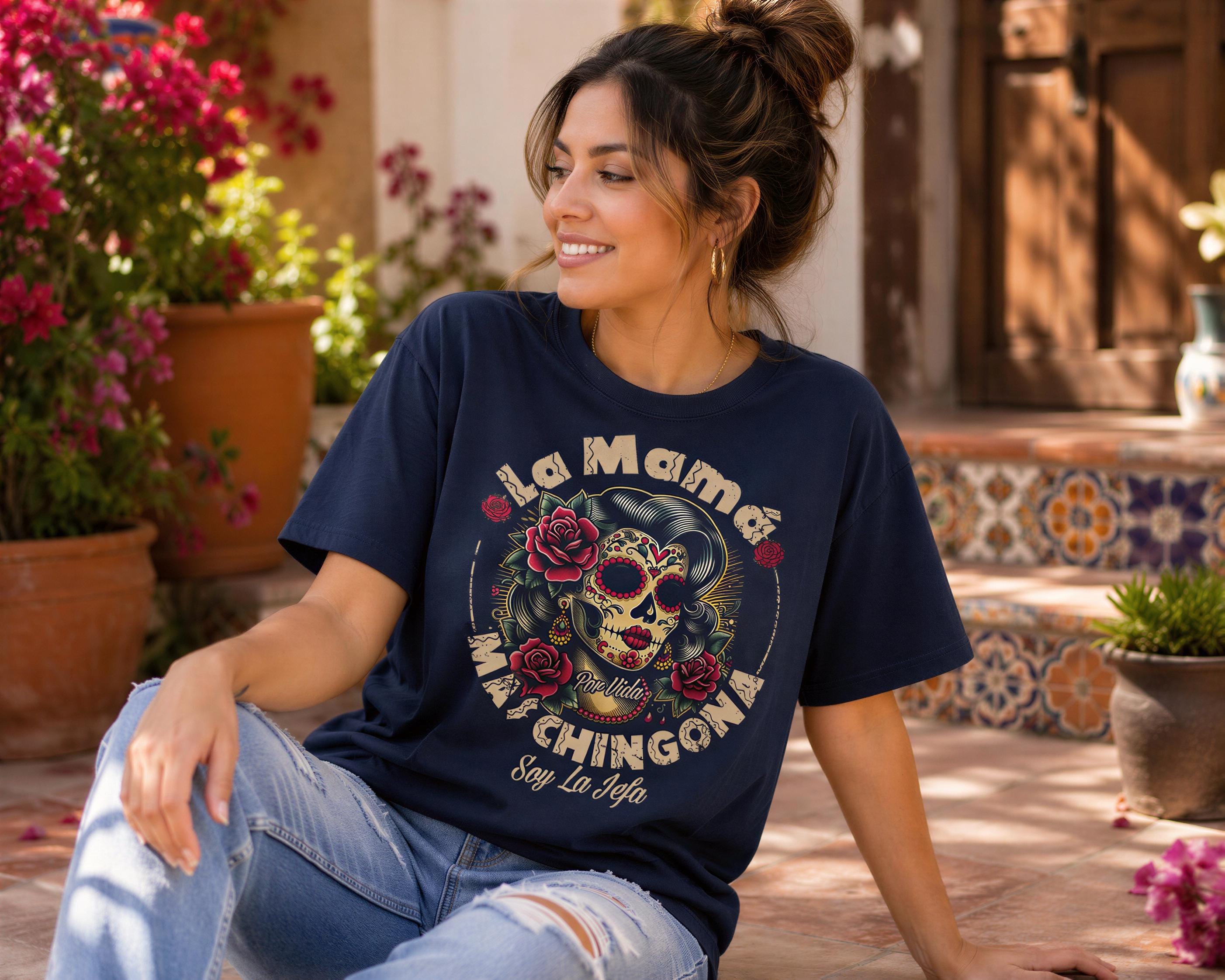 A woman wearing a blue t-shirt with a graphic design of a skull and roses, sitting on a patio surrounded by potted plants and flowers.