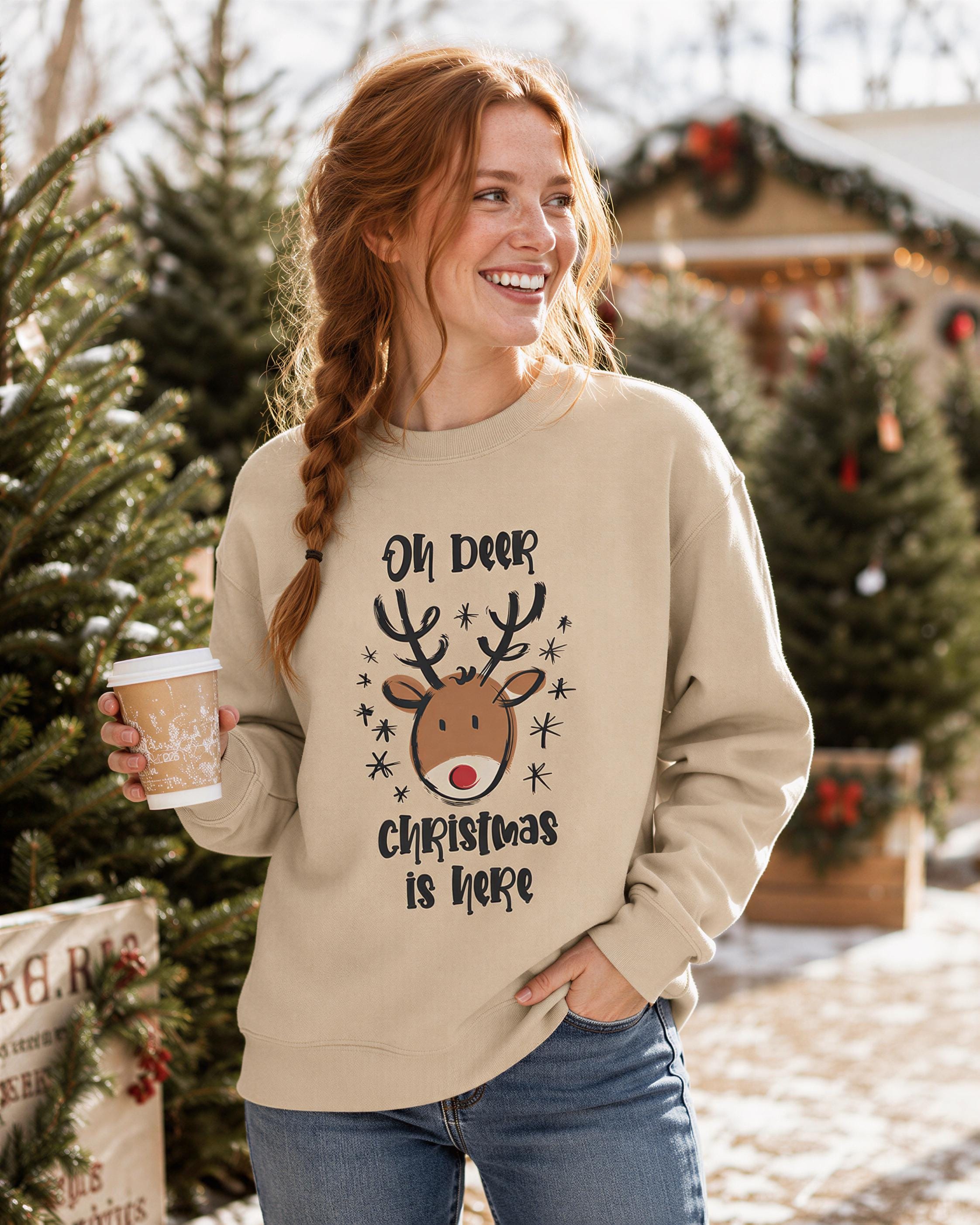 A woman wearing a beige sweater with a Christmas-themed design and holding a coffee cup, smiling and standing in front of Christmas trees.