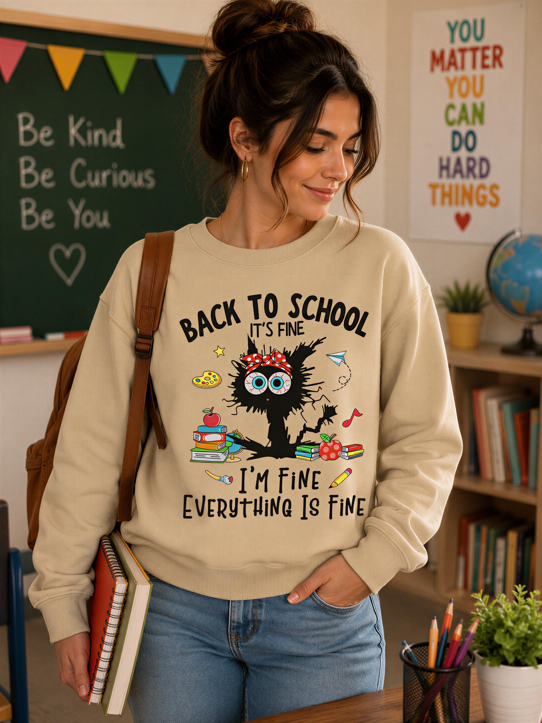 A woman wearing a beige sweatshirt with a cat graphic and text, standing in a classroom setting with a chalkboard and bookshelves in the background.