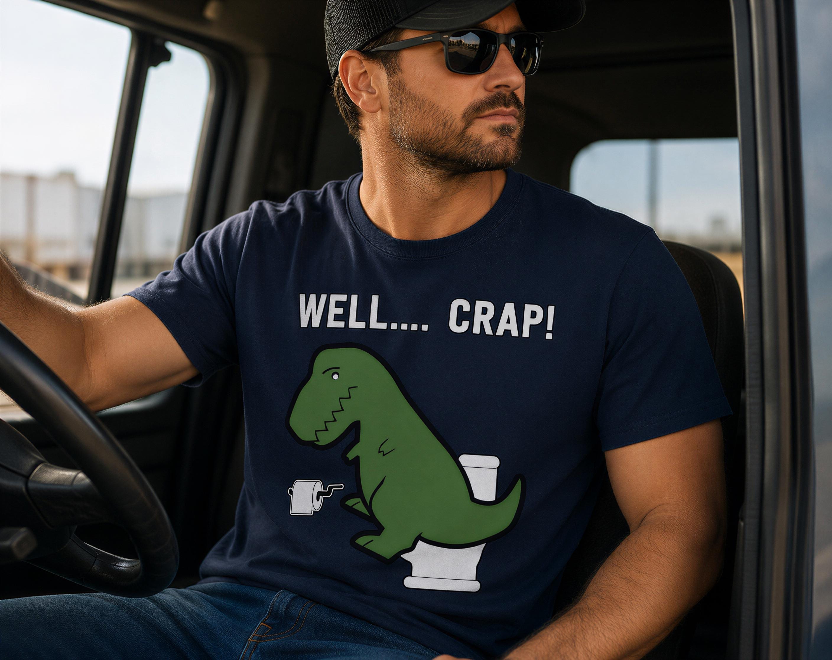 A man wearing a blue t-shirt with a graphic of a green dinosaur and the text "WELL... CRAP!" is sitting in a vehicle.