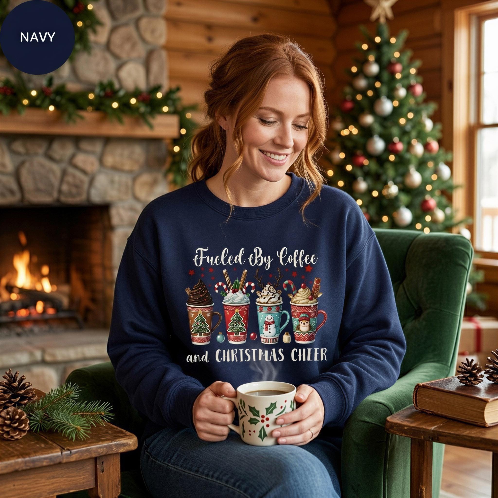 A woman wearing a navy blue sweatshirt with a Christmas-themed design is sitting in a cozy living room, holding a mug of coffee and smiling.
