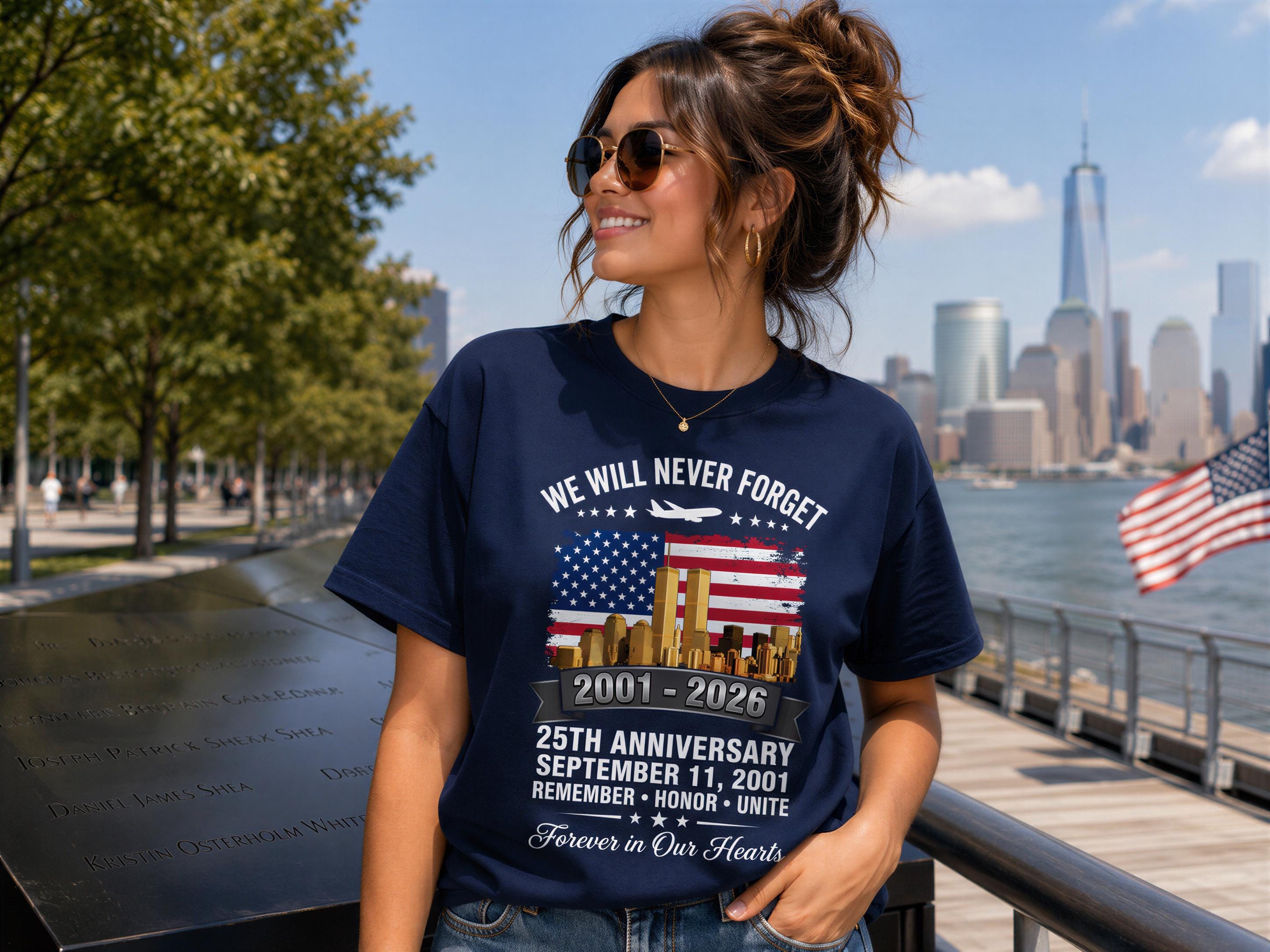 A woman wearing a navy blue t-shirt with a graphic design celebrating the 25th anniversary of September 11, 2001, stands in front of a memorial with the American flag and the New York City skyline in the background.