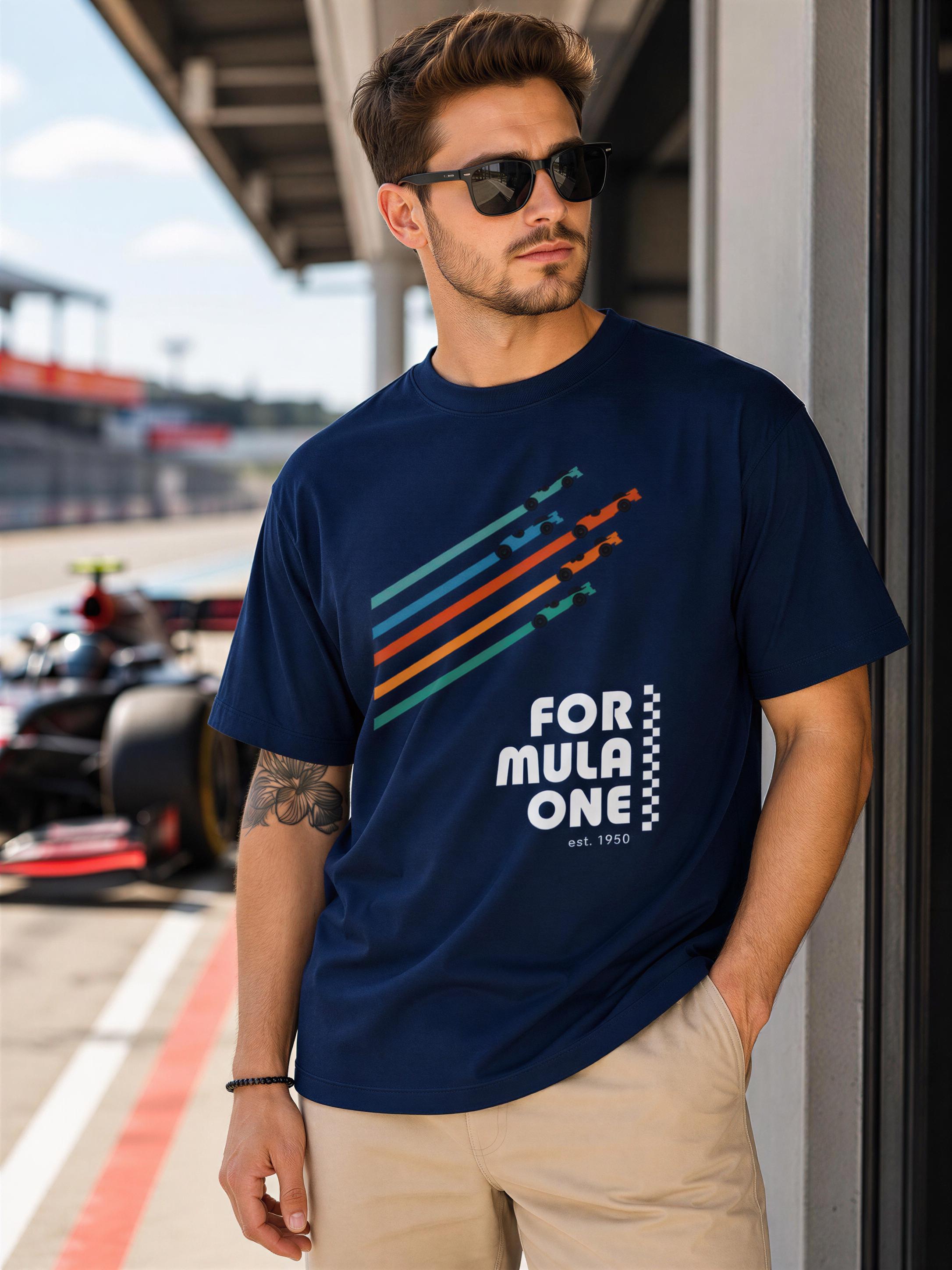 A man wearing sunglasses and a navy blue t-shirt with a graphic design stands in front of a race track with a Formula One car.