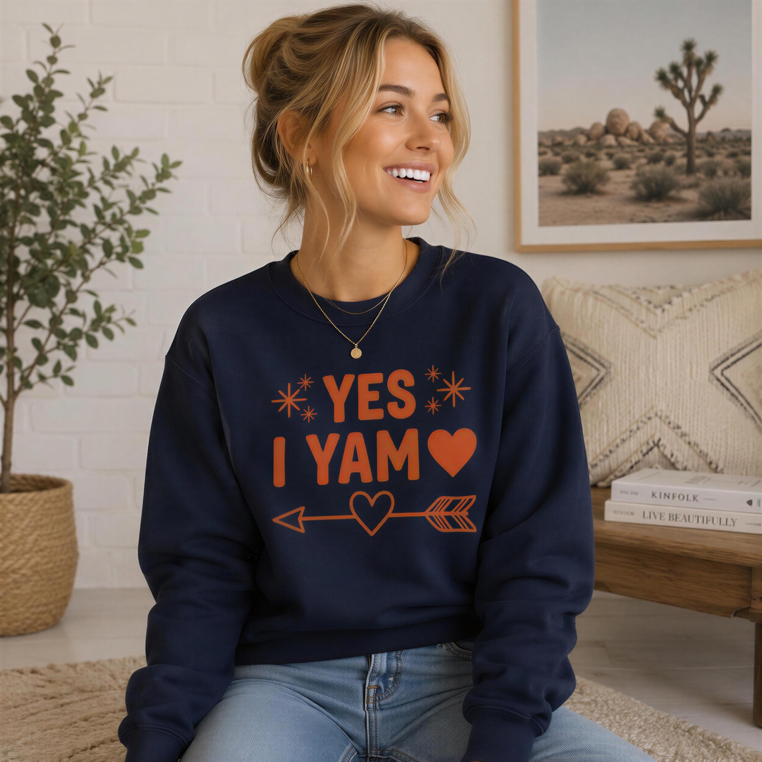 A smiling woman wearing a navy blue sweatshirt with the text "YES I YAM" and an arrow graphic, sitting on a couch in a room with a potted plant and a framed picture of a desert landscape.